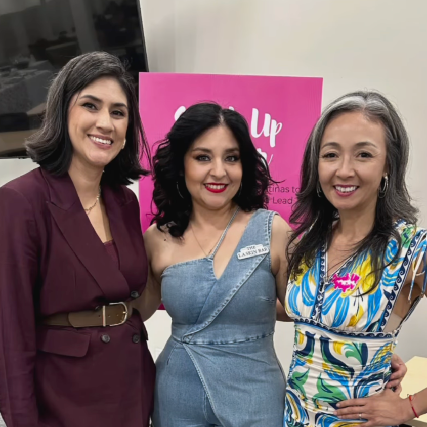 Three women smiling and standing together indoors, with a pink sign behind them. The woman on the left wears a burgundy blazer, the woman in the middle wears a denim dress, and the woman on the right wears a colorful patterned dress.