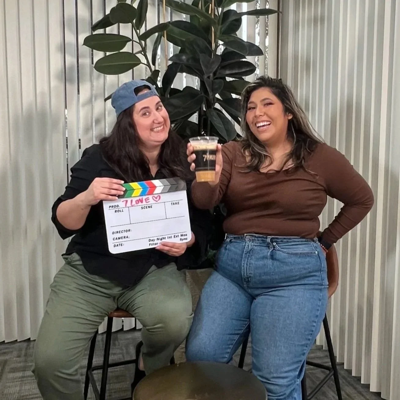 Two women smiling, one holding a film slate, the other holding a pint glass of beer, sitting in front of a large plant with vertical blinds in the background.