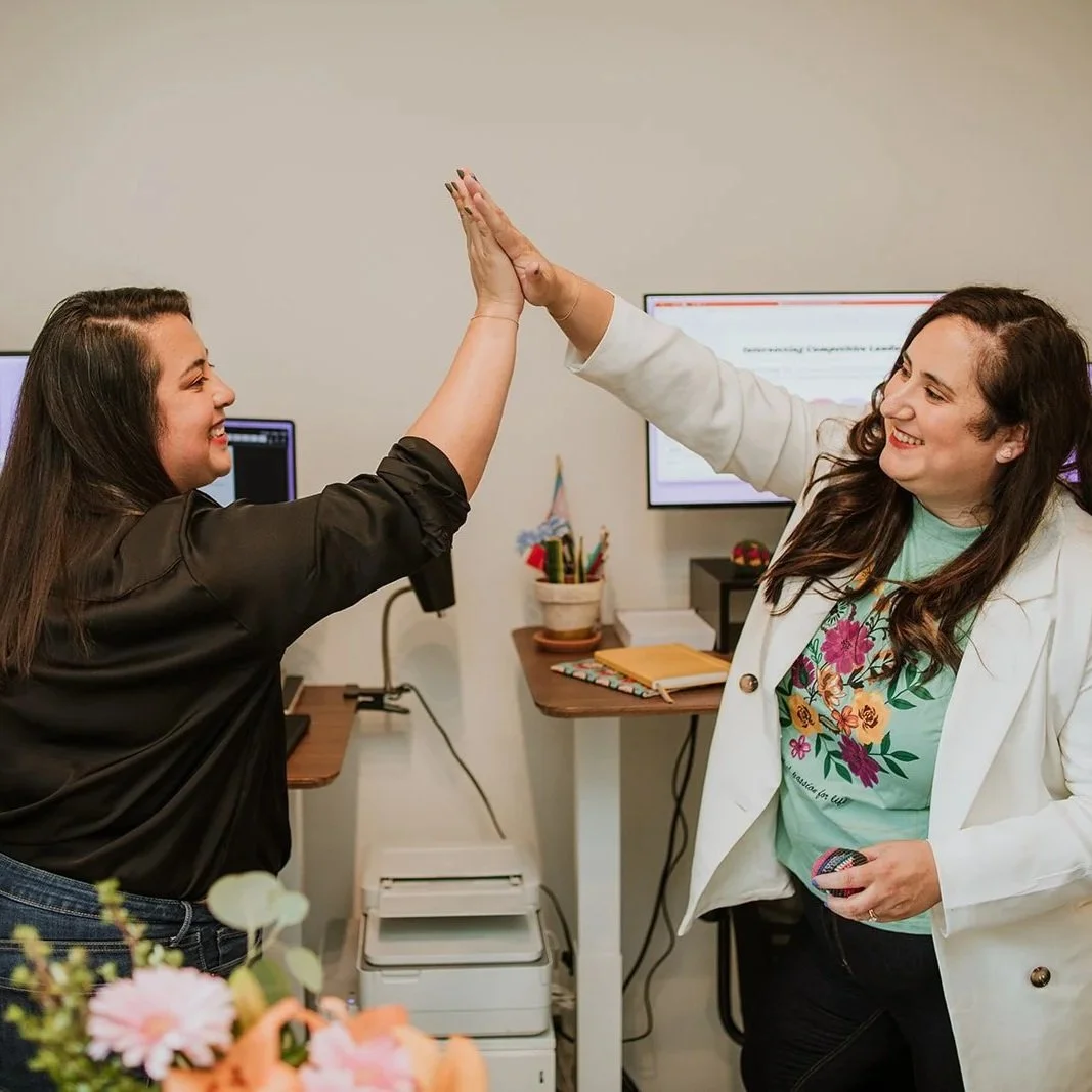 Two women giving each other a high five in an office, smiling. One is wearing a black shirt, the other a white blazer with a floral shirt underneath. There are computer monitors, a printer, and office supplies in the background.