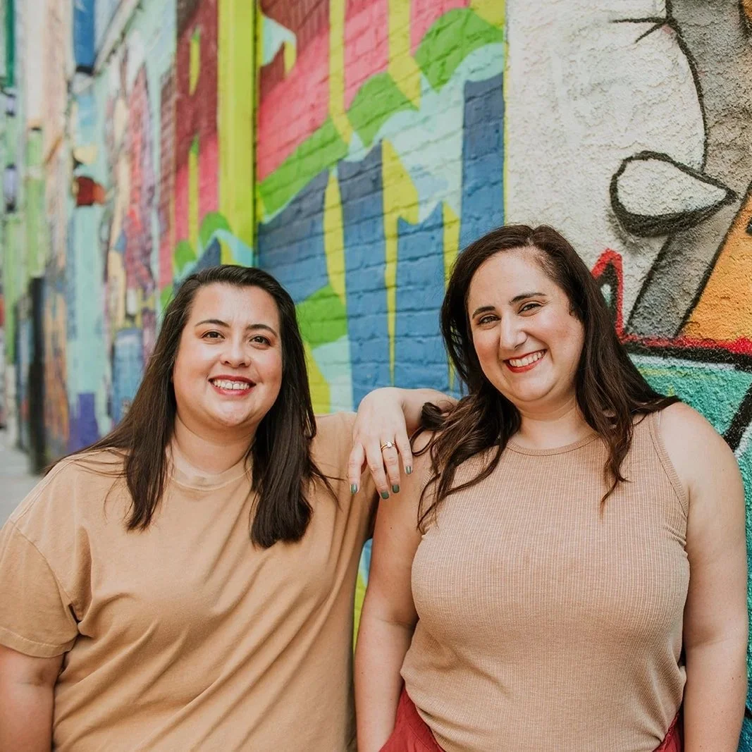 Two women smiling, standing in front of a colorful graffiti wall, one with her arm around the other's shoulder.