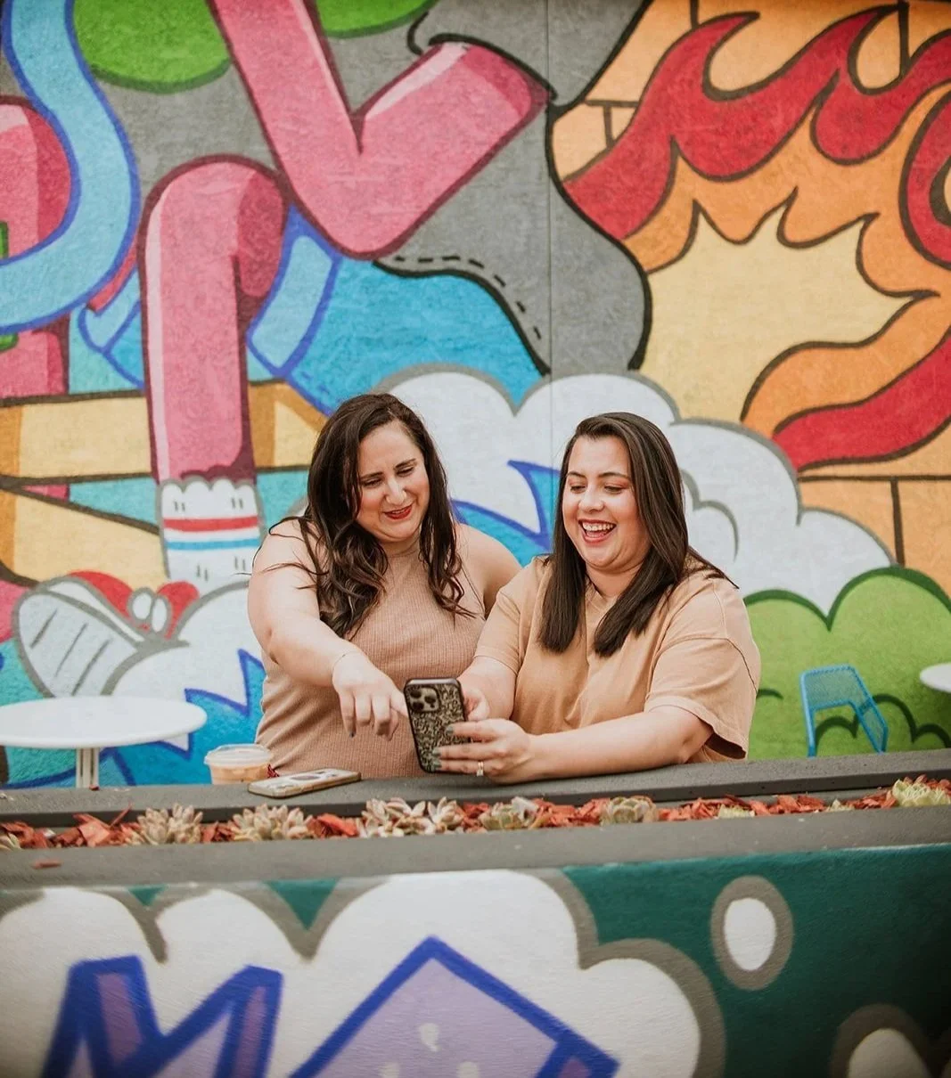 Two female adults standing next to each other, one holding a phone & the other woman pointing at the device. They both have brown hair and nude colored shirts. Behind them is a vibrant mural in a cartoon style.