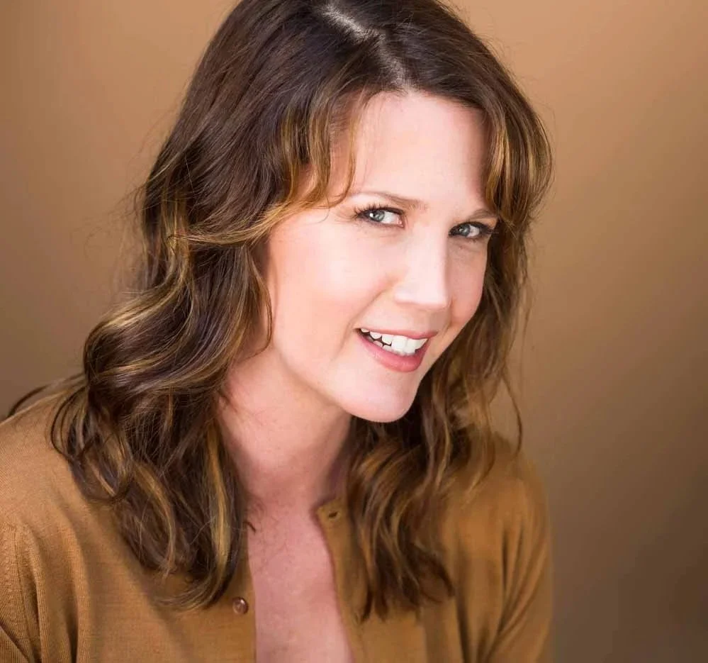 Adria, speaking coach for excutives ,with wavy, shoulder-length brown hair with highlights, wearing a brown top, smiling, and looking slightly to her right, with a neutral background.