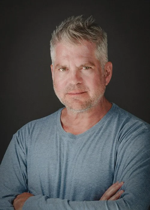 A middle-aged man with gray hair and a beard, wearing a light blue shirt, looking at the camera with a neutral expression, against a dark background.