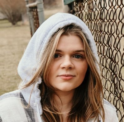 Young woman with long hair wearing a hoodie, standing outdoors next to a chain-link fence.