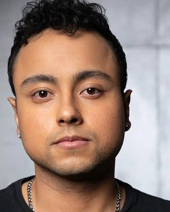 Close-up portrait of a young man with short curly hair, earrings, and a chain necklace, against a gray background.