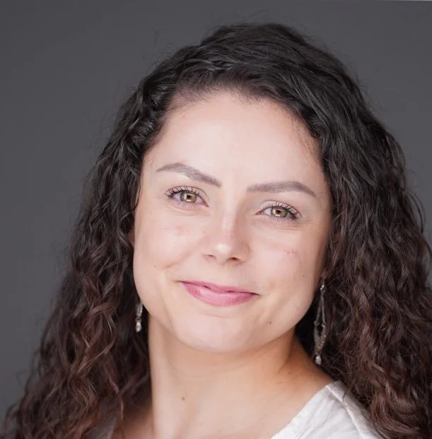 Close-up portrait of a woman with dark, curly hair, light skin, and a gentle smile, wearing earrings against a gray background.