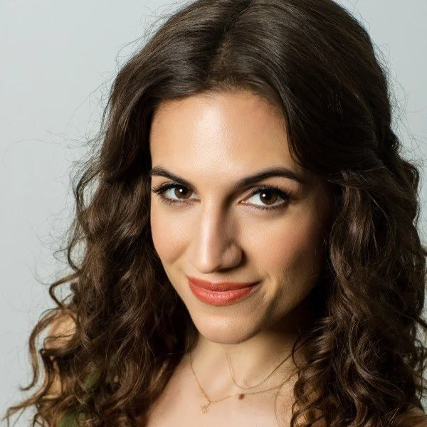 Close-up of a woman with long, curly brown hair, wearing subtle makeup and a delicate gold necklace, smiling softly at the camera.
