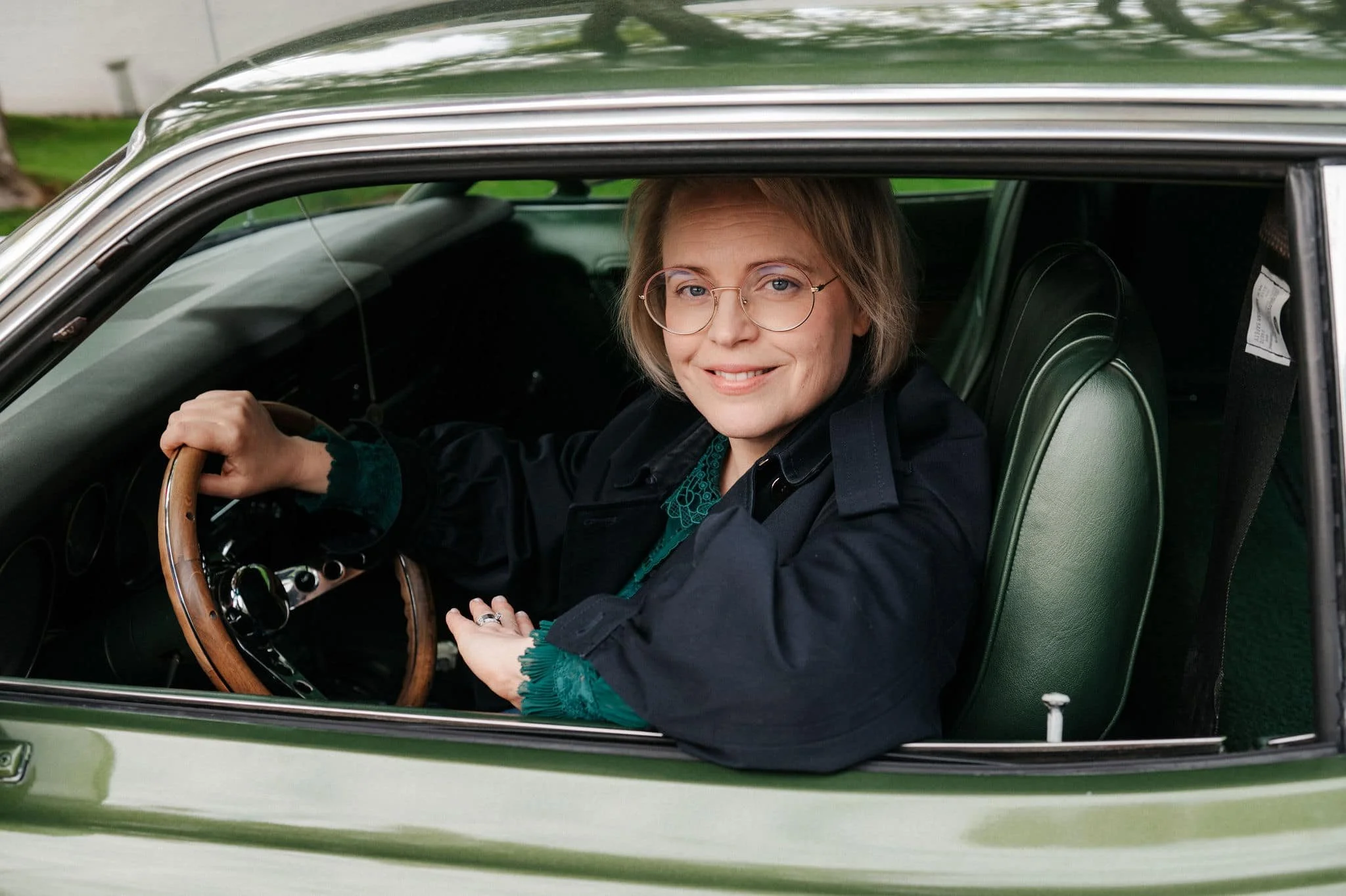 Nainen istuu auton kuljettajan paikalla, hymyilee, ja katsoo kameraan, hänellä on silmälasit ja lyhyt vaaleanruskea tukka.