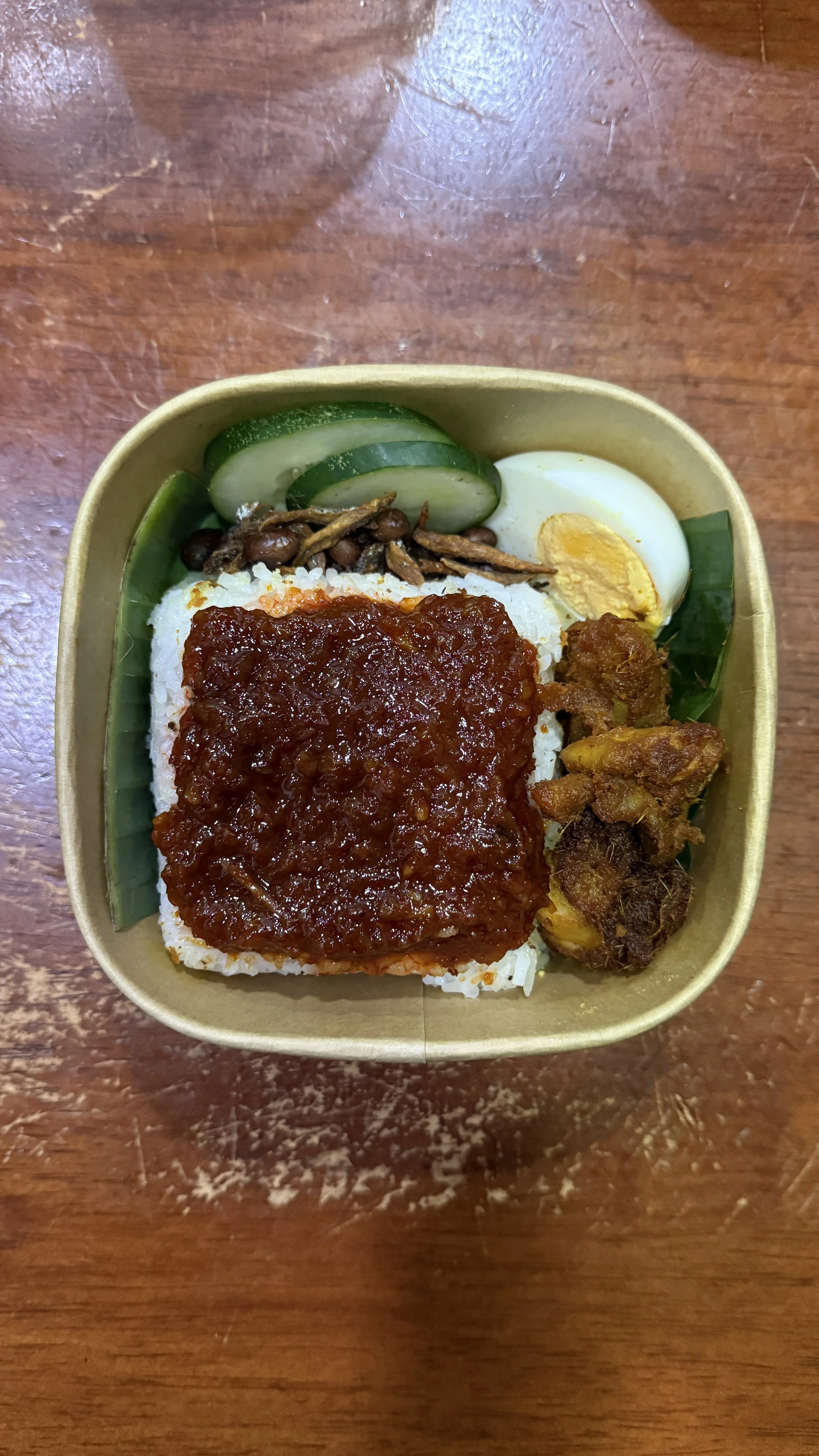 A bento box with white rice topped with a dark brown sauce, cucumber slices, a halved boiled egg, and some cooked vegetables and chicken on a wooden table.