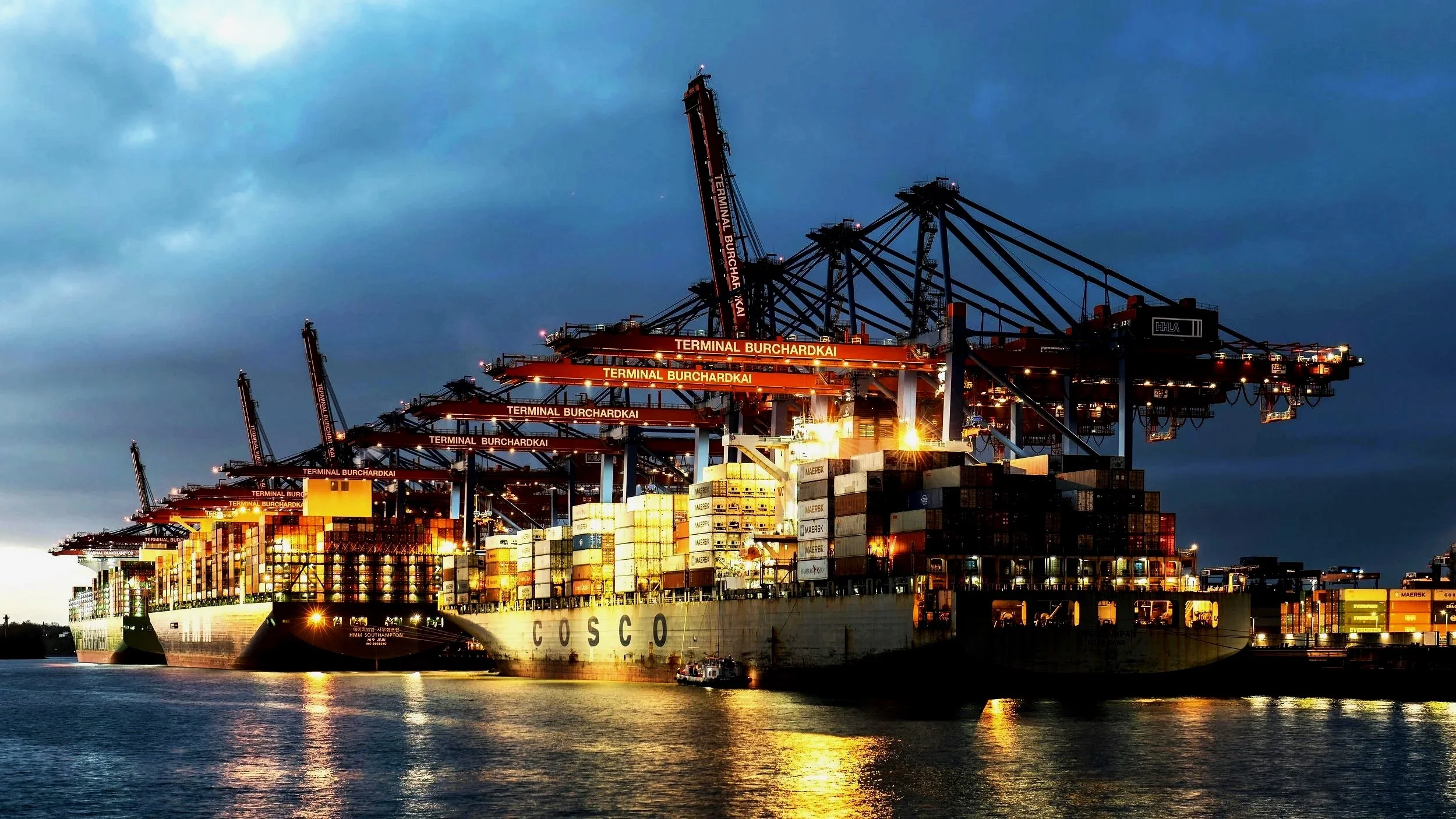 A large cargo ship docked at a port with numerous cranes and containers, illuminated at dusk with a dark sky overhead.