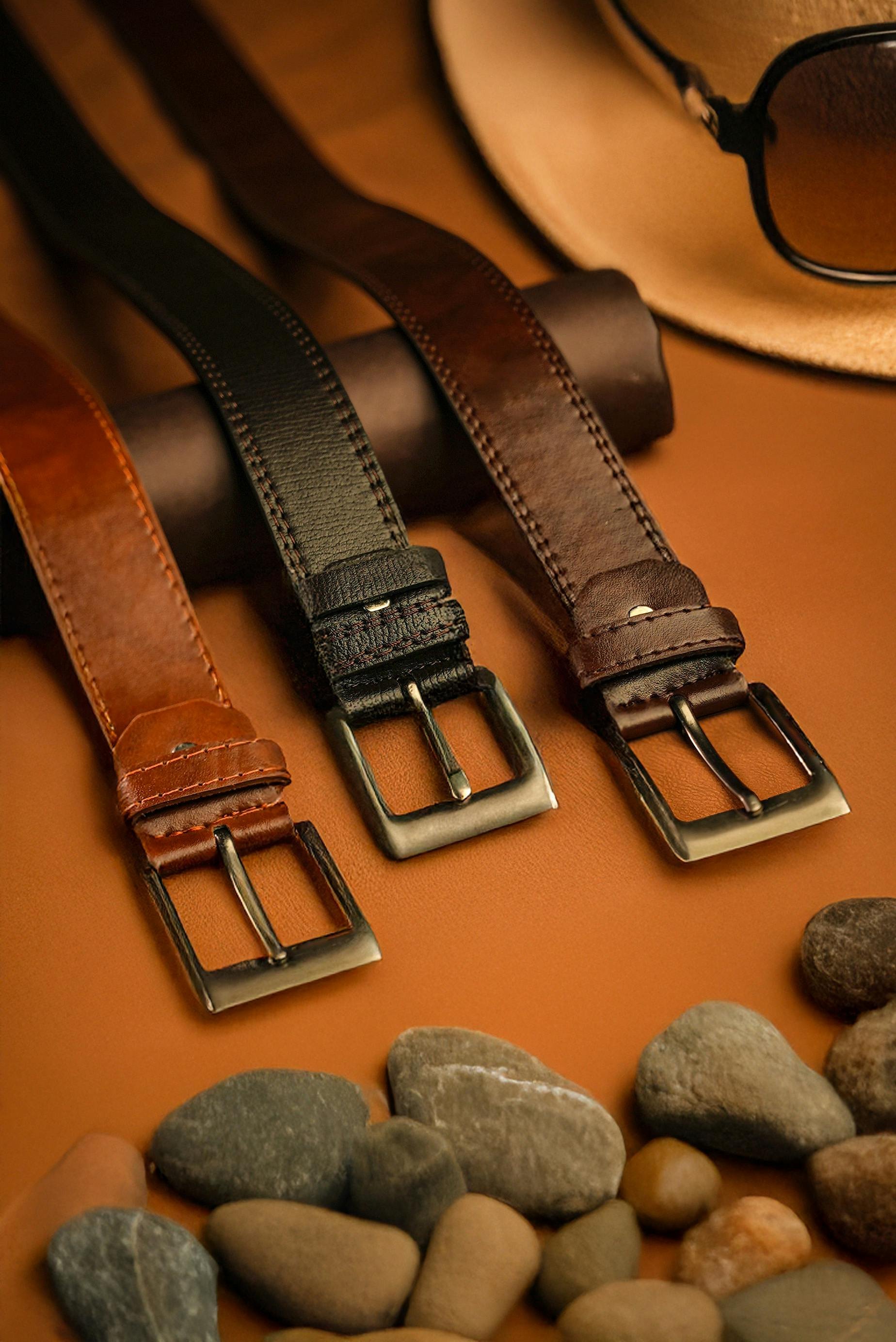 Three leather belts in brown, black, and dark brown colors laid out on a surface with small rocks at the bottom and a fedora hat with sunglasses in the background.