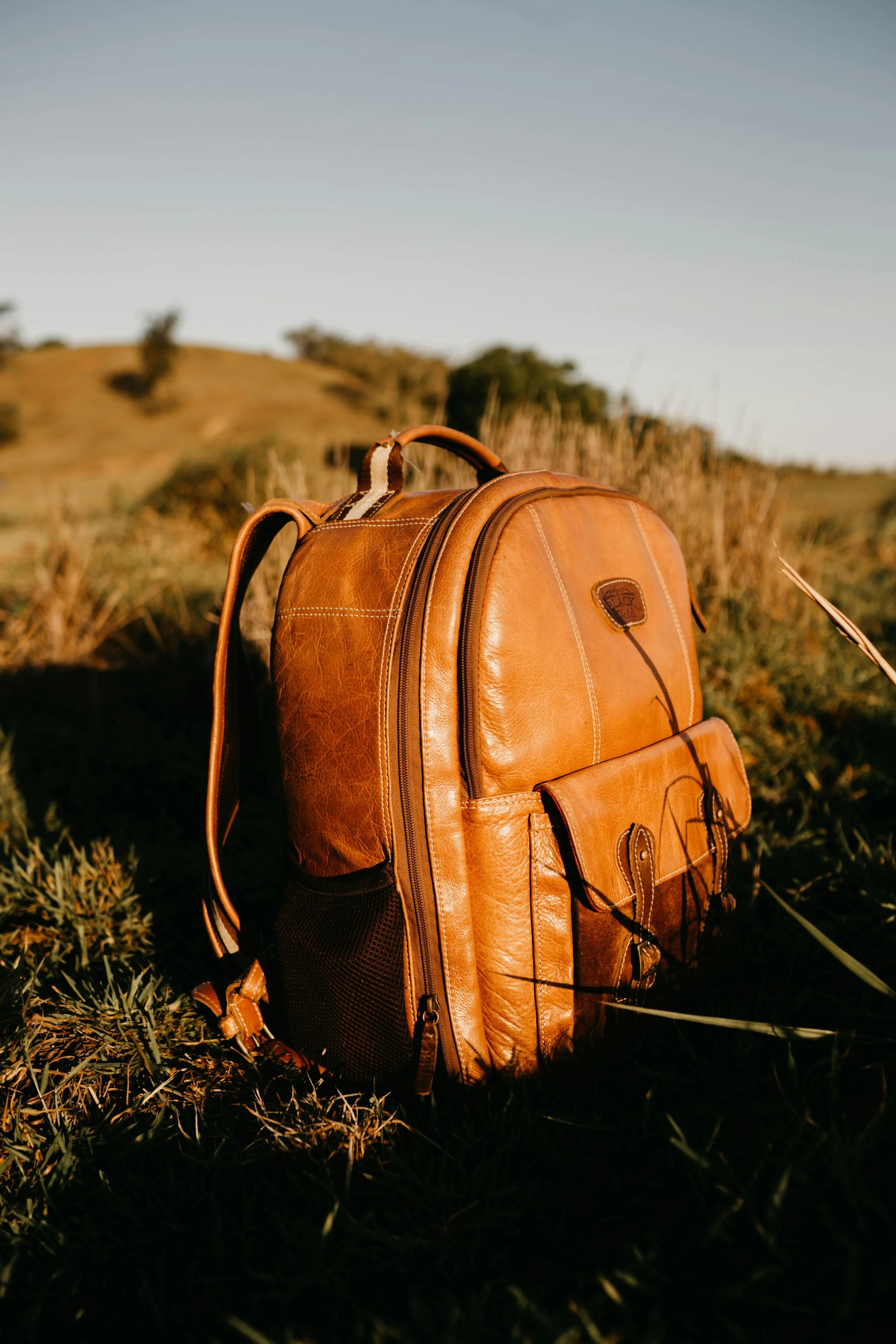 A brown leather backpack resting on green grass during sunset, with a hilly landscape in the background.