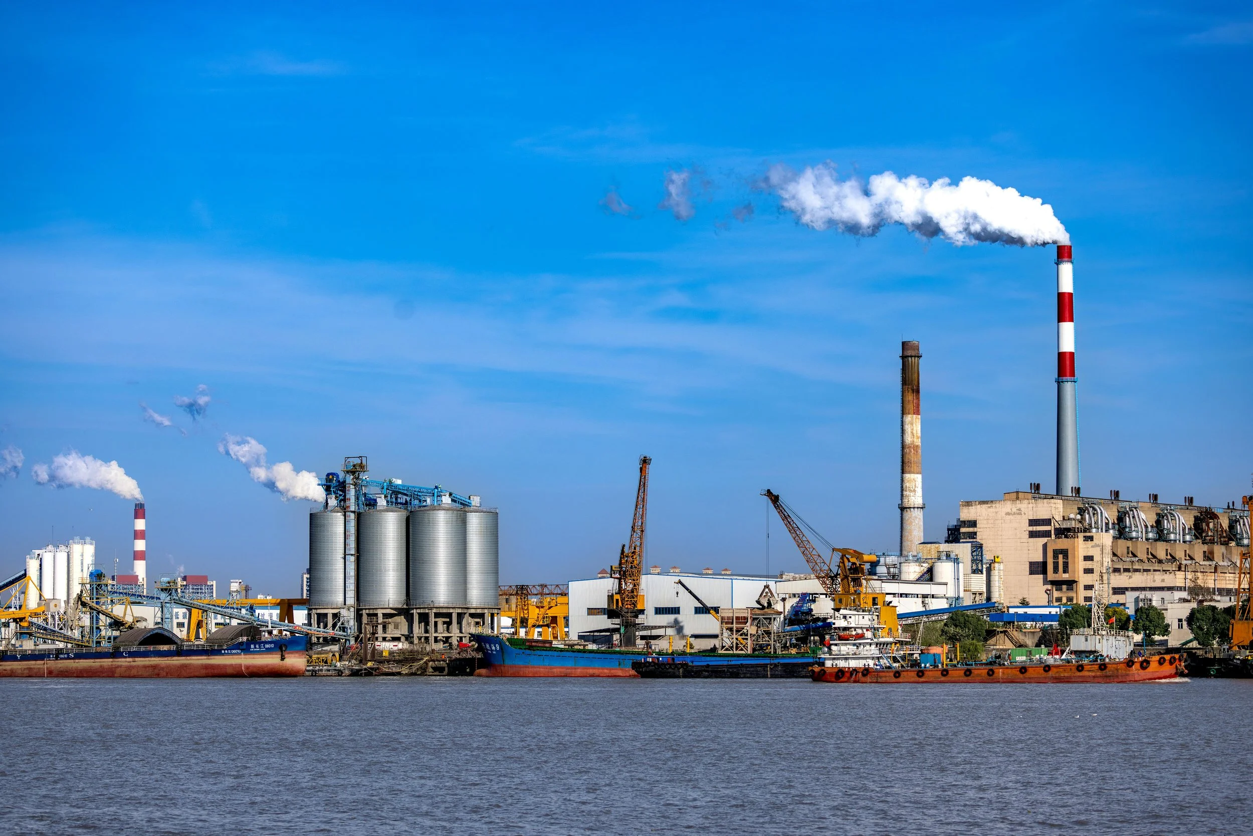 Industrial factory with multiple smokestacks emitting white smoke, large tanks, cranes, ships, and warehouses along a waterfront under a blue sky.