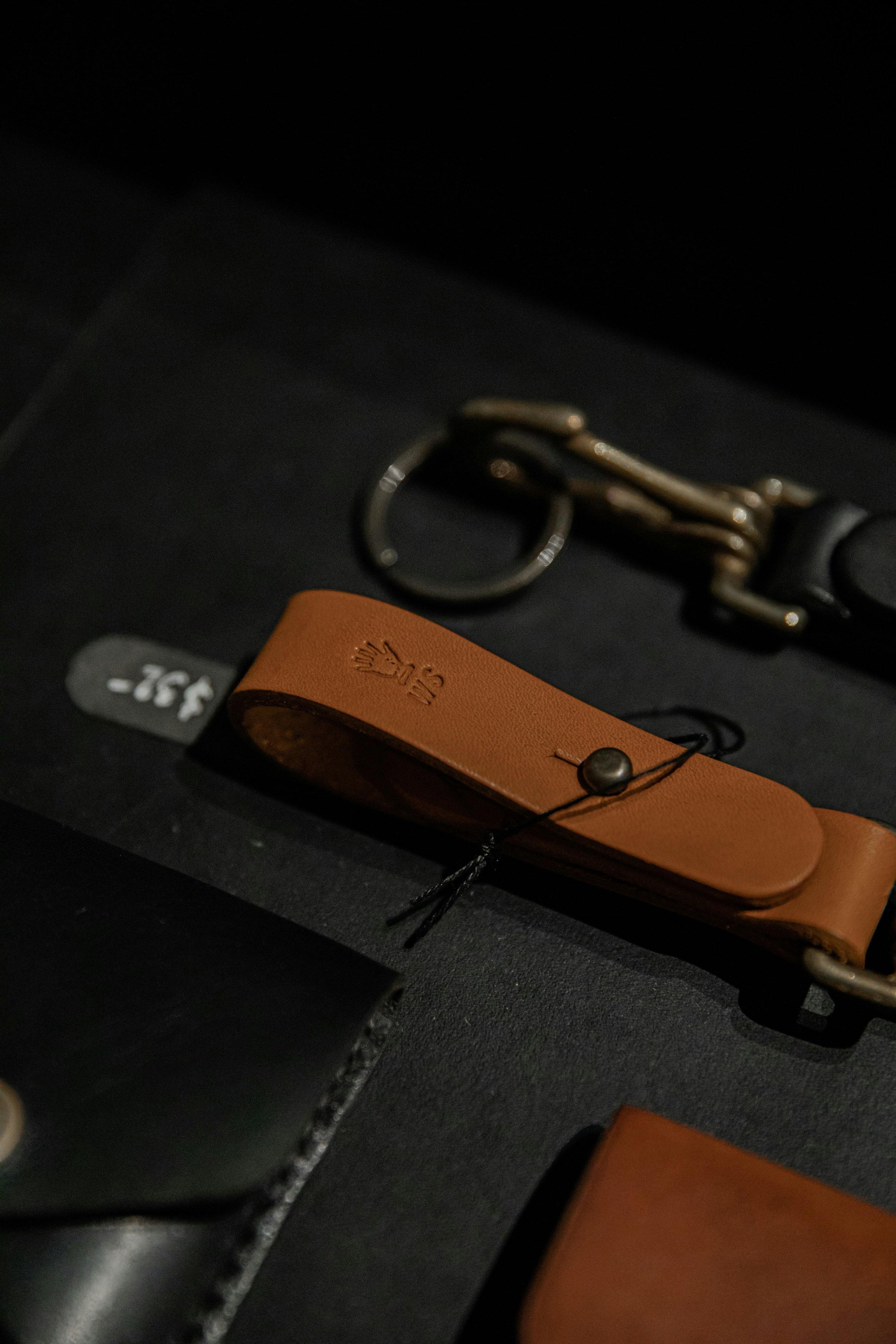 Close-up of leather accessories, including a tan leather strap with embossed logo, a keyring, and metal hardware, on a dark surface.