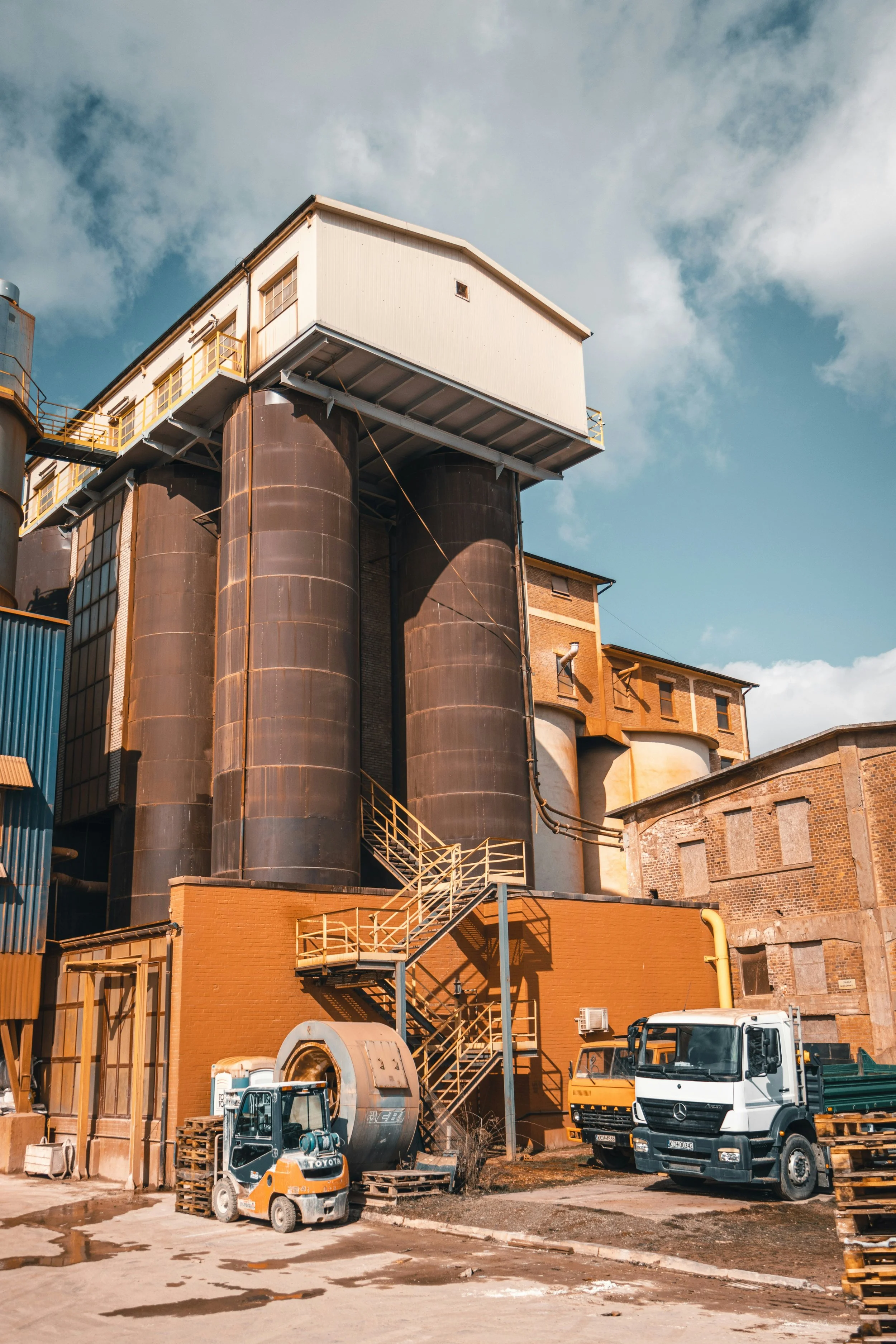 Old industrial building with large black silos, trucks, forklift, and staircases, under a cloudy sky.