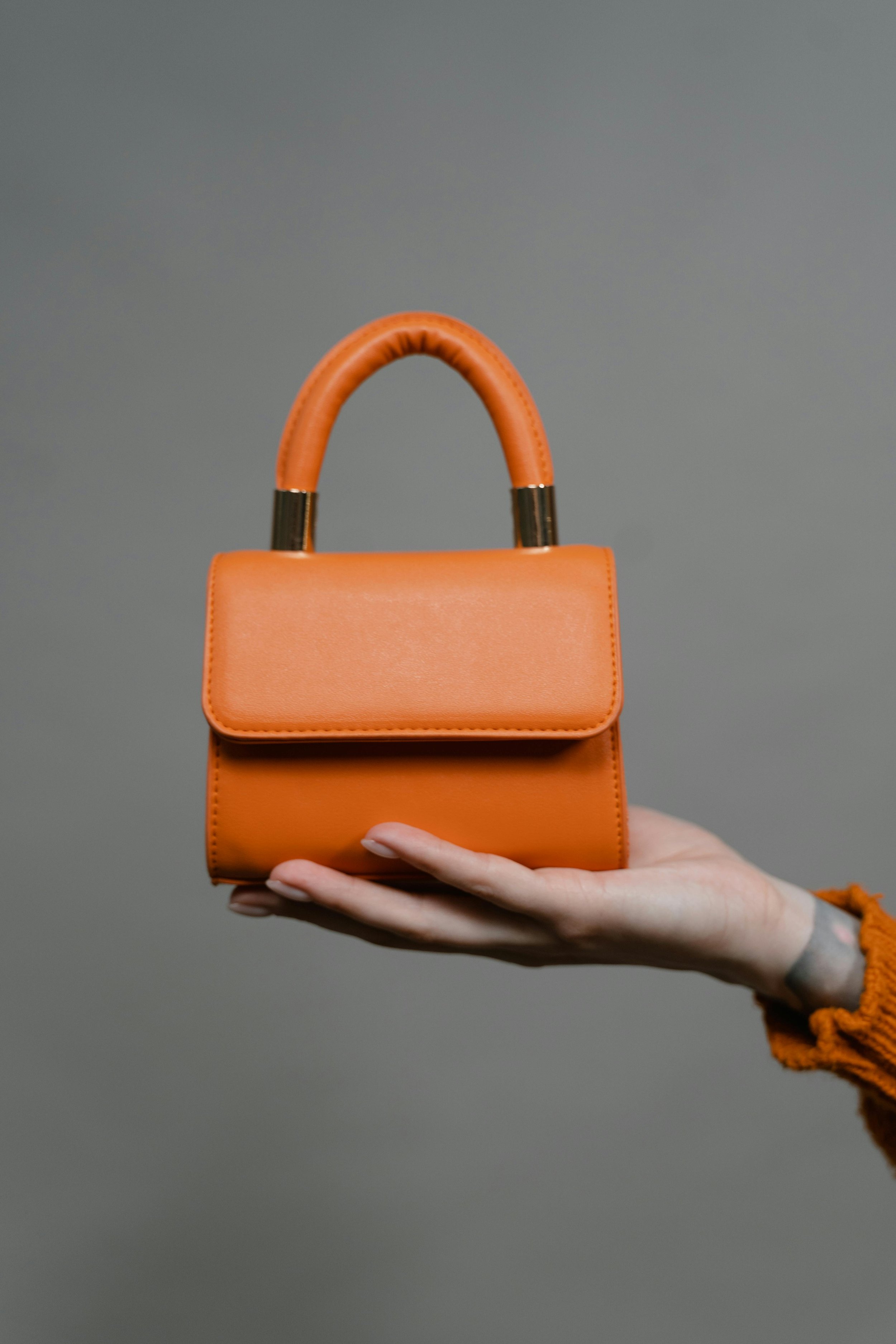 A small, orange handbag with a rounded handle, held by a person's hand against a plain, gray background.