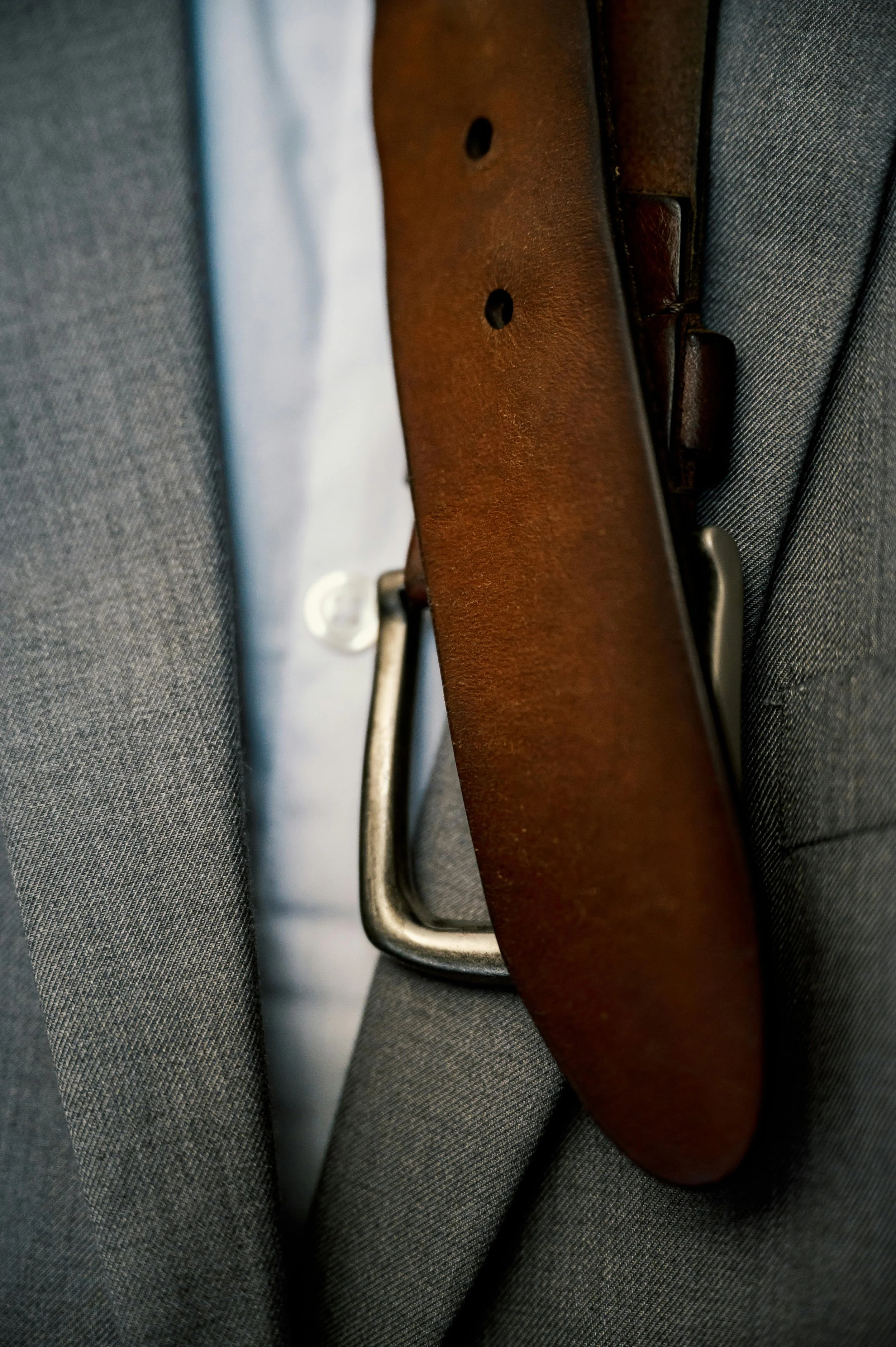 Close-up of a gray suit, white shirt, and brown leather belt with a silver buckle.
