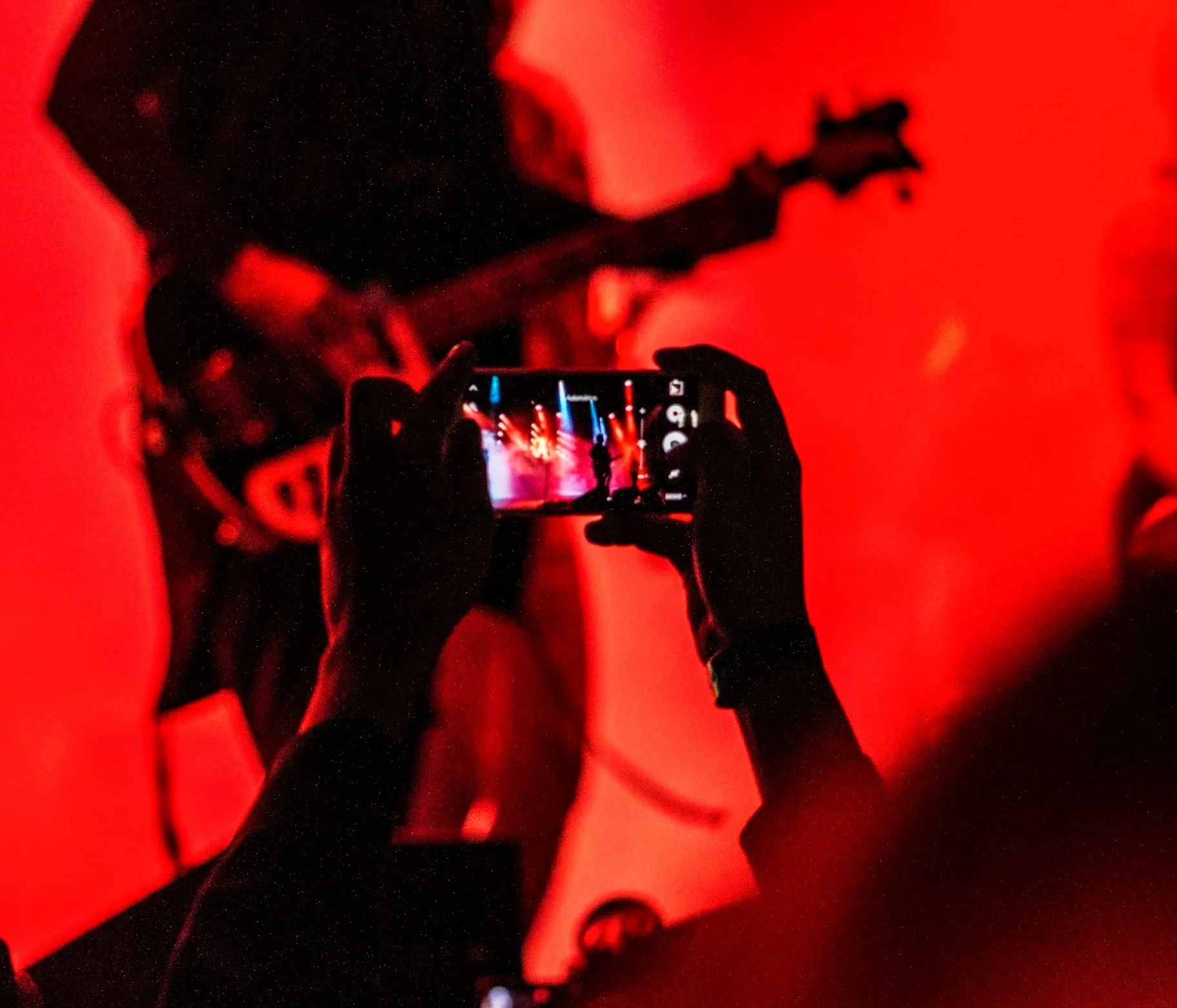 Person taking a photo of a stage performance with a smartphone, visible stage lights, musicians, and a microphone on stage, all in red lighting.