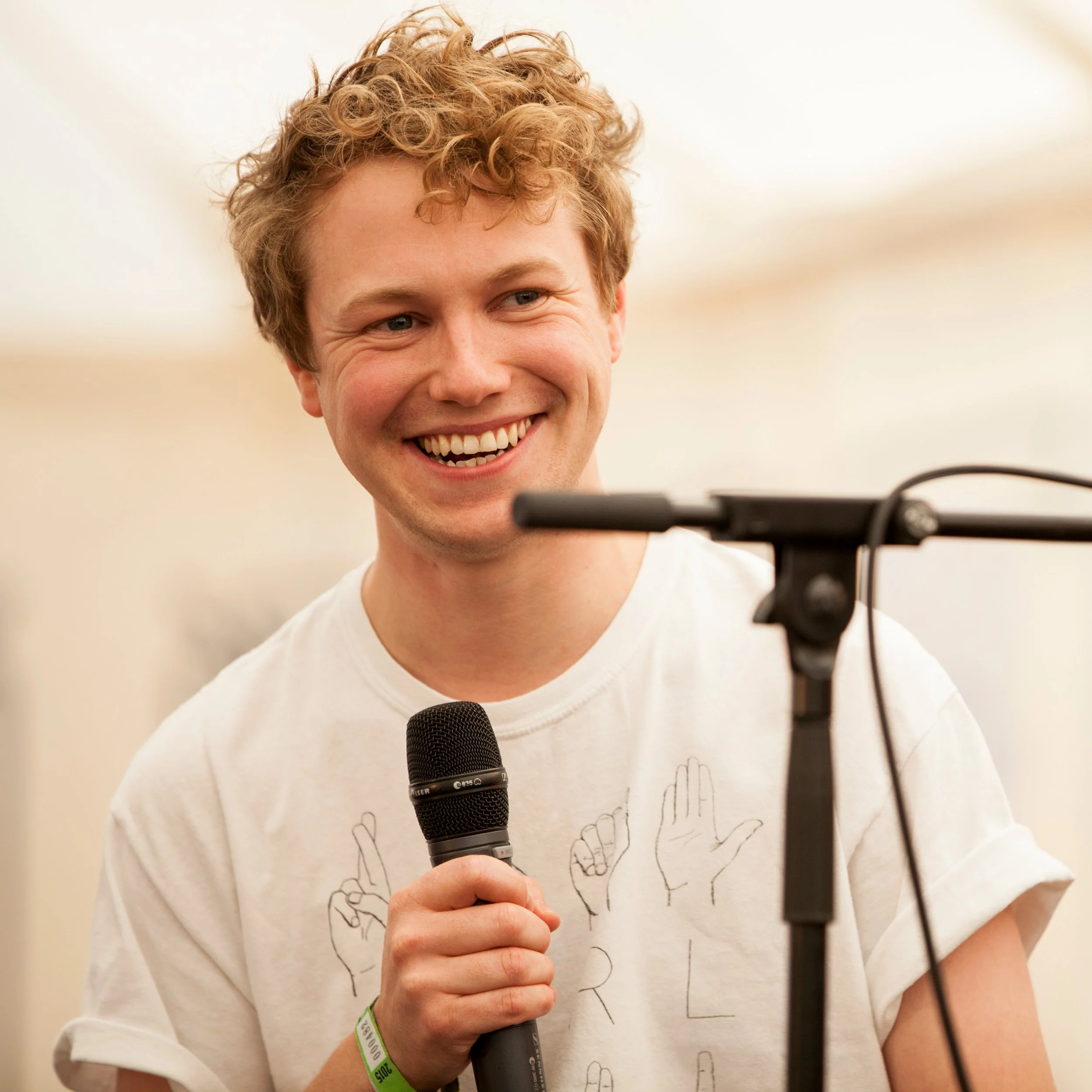A young man with curly hair smiling while holding a microphone in front of a microphone stand.