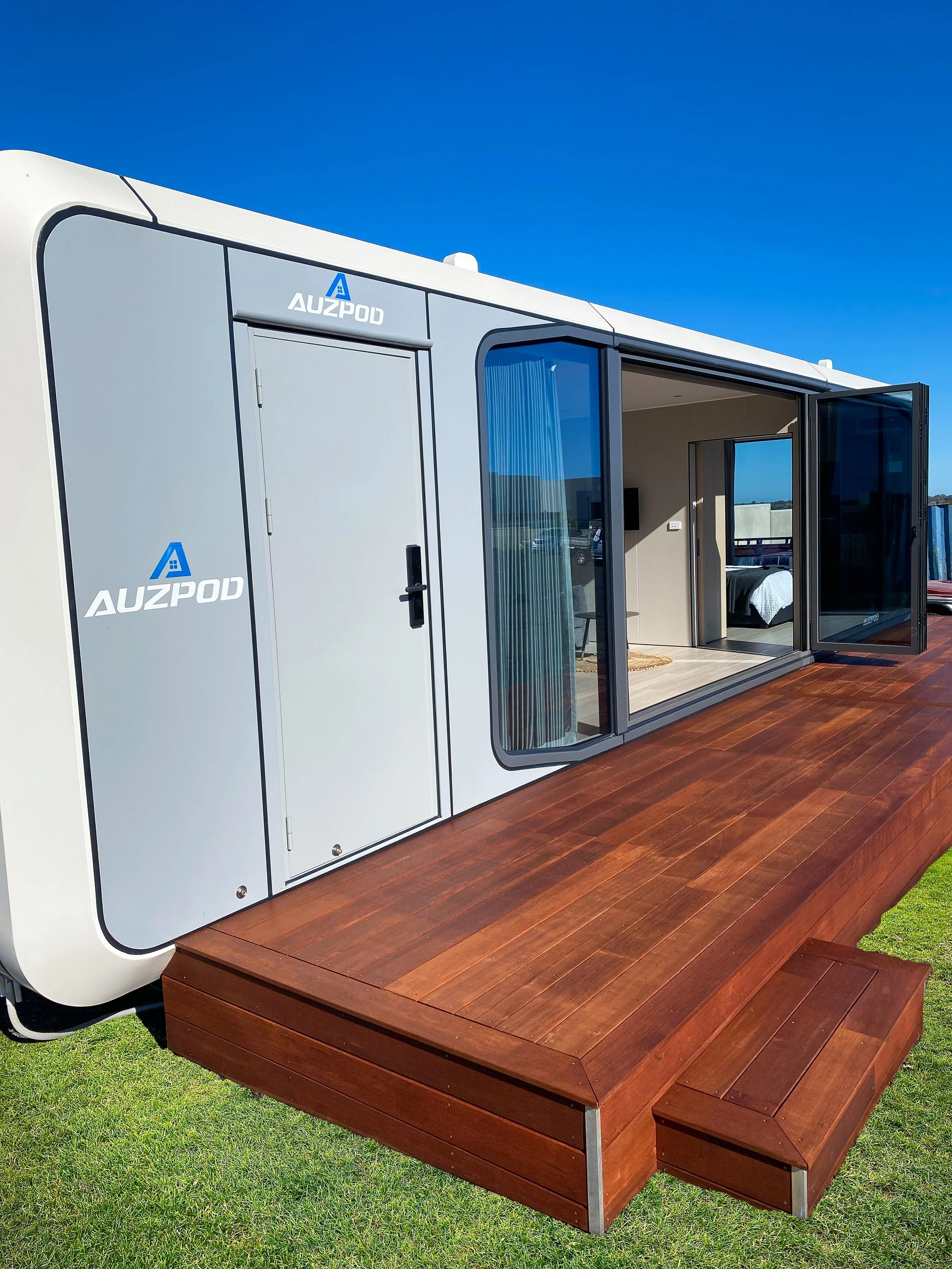 AUZPOD sustainable modular pod house installed on grass with timber decking, sliding doors open to reveal the interior living space.