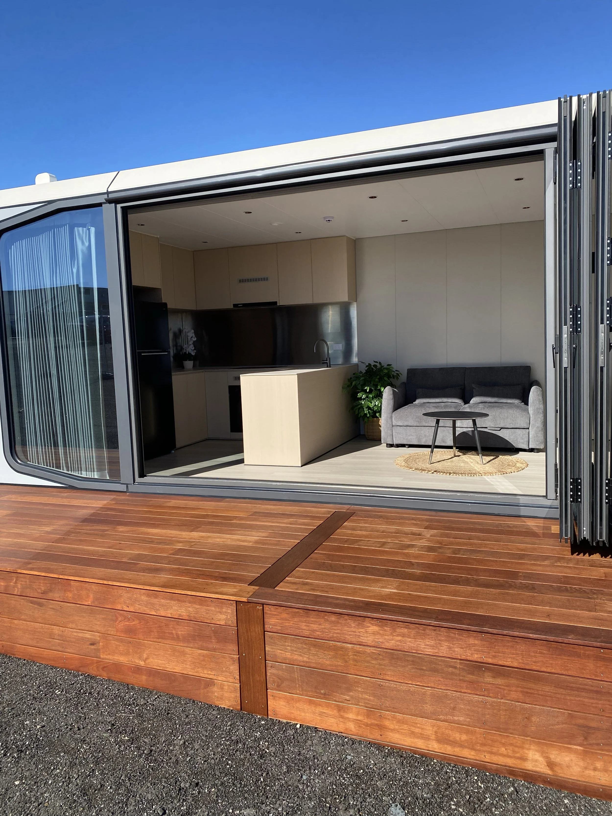 Interior of a modern AUZPOD sustainable pod house featuring a kitchen, grey sofa, small black coffee table, potted plant, and large sliding glass doors opening to a wooden deck.
