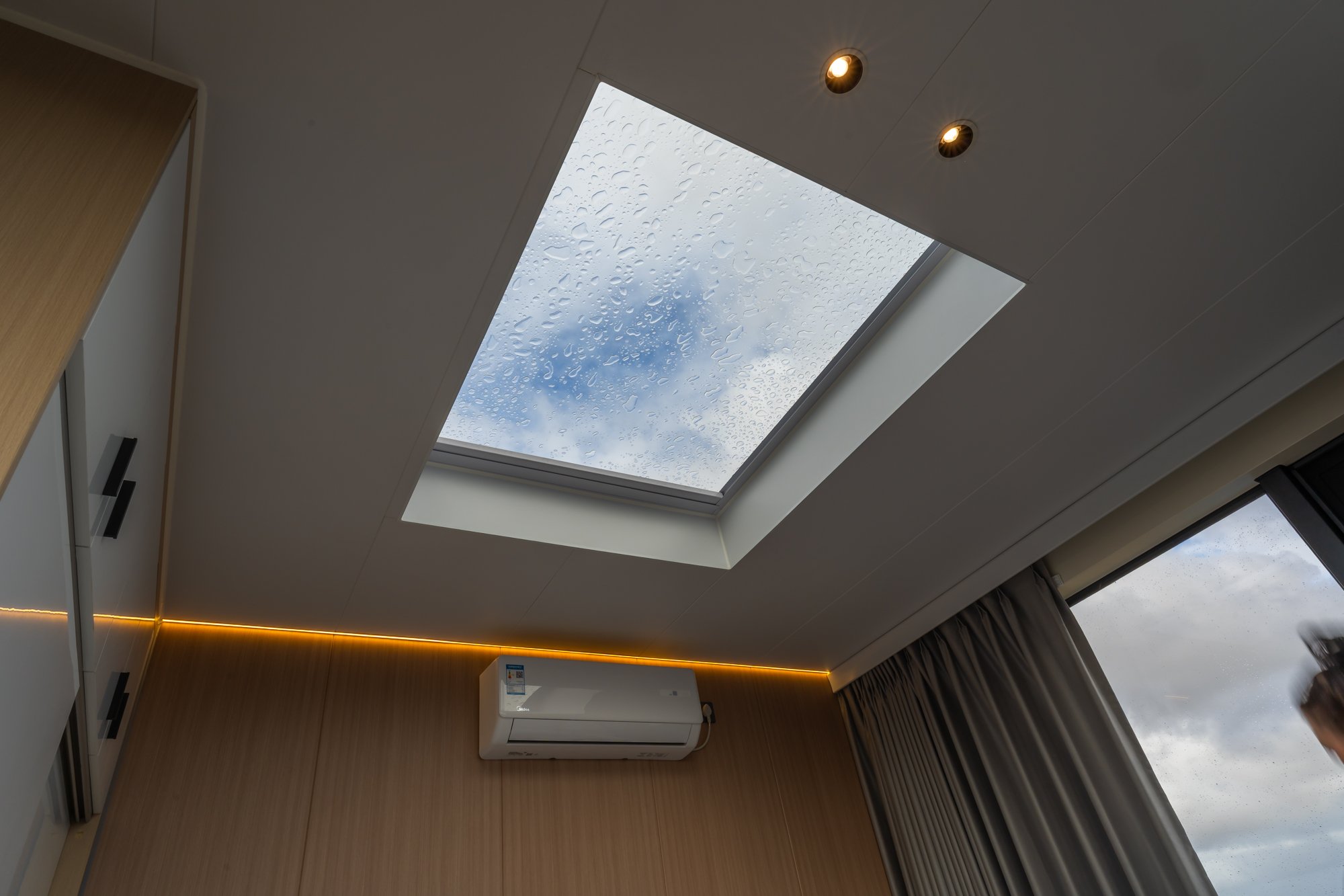 AUZPOD sustainable modular pod house bedroom ceiling with skylight showing raindrops, spotlights, air conditioning unit, and window curtains.