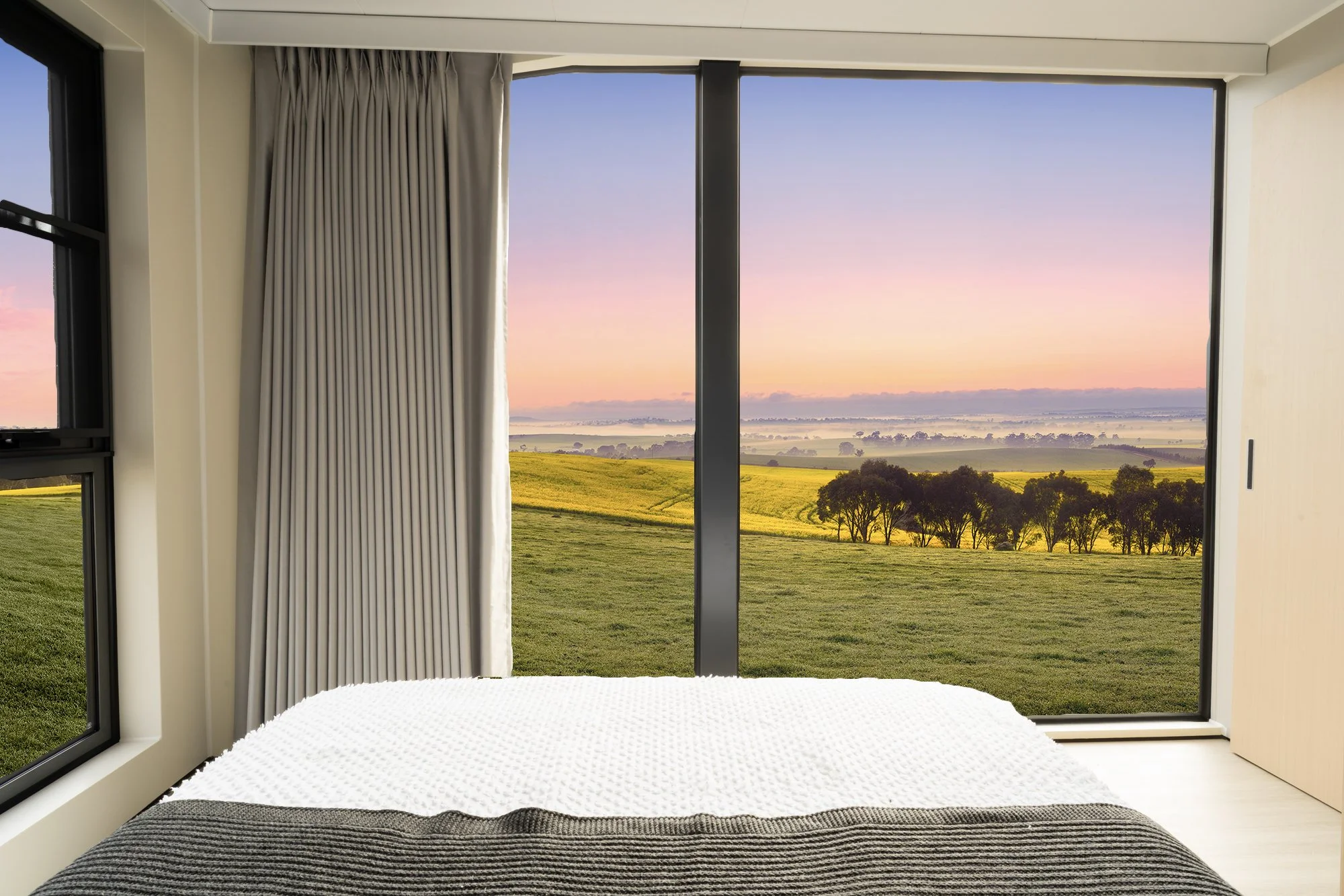 AUZPOD sustainable modular pod house bedroom with large glass sliding door opening to scenic countryside view at sunset, overlooking fields, trees, and a colourful sky.