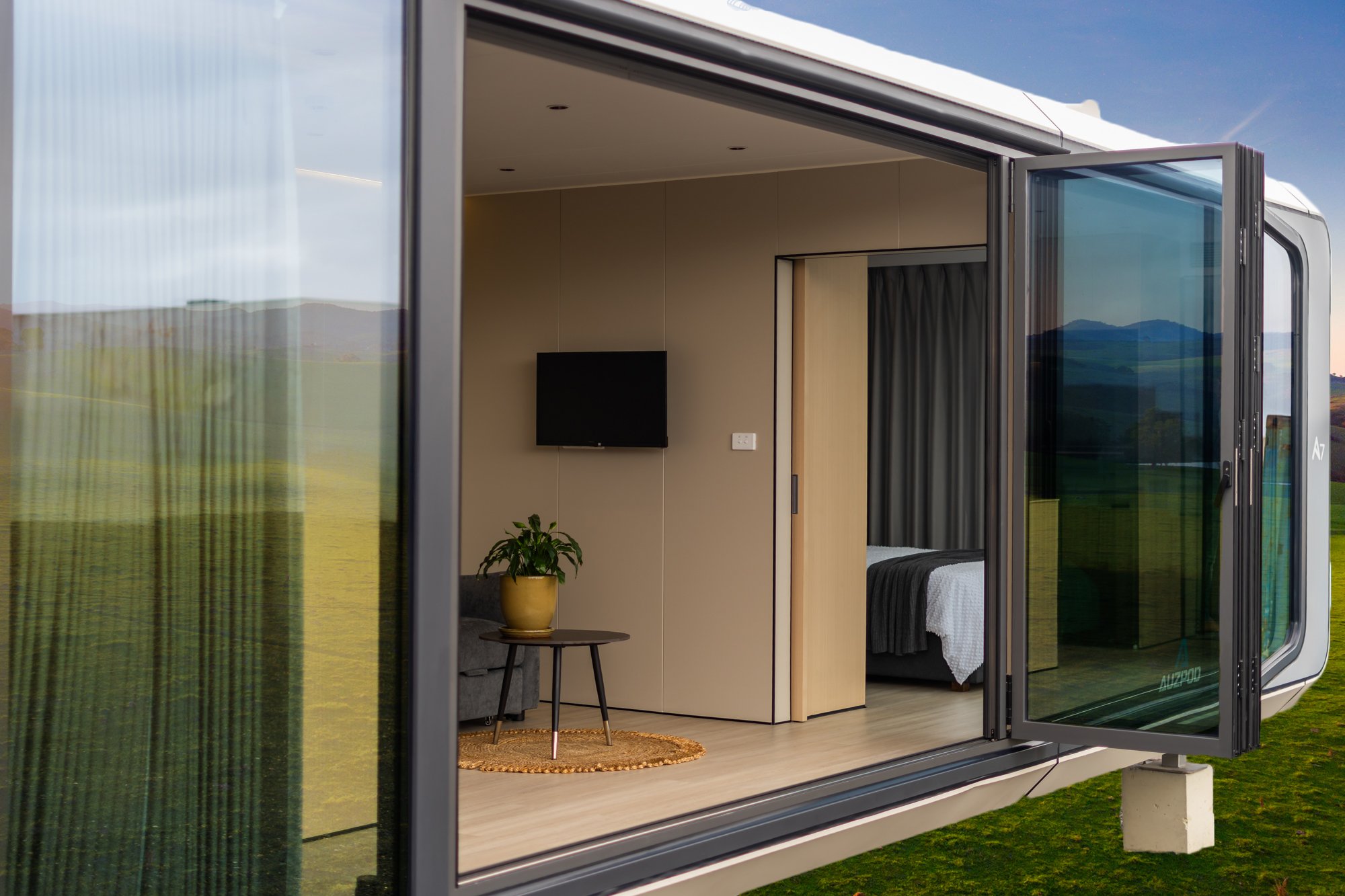 AUZPOD sustainable tiny pod house interior with wall-mounted TV, potted plant, small table, and bed visible through open window.