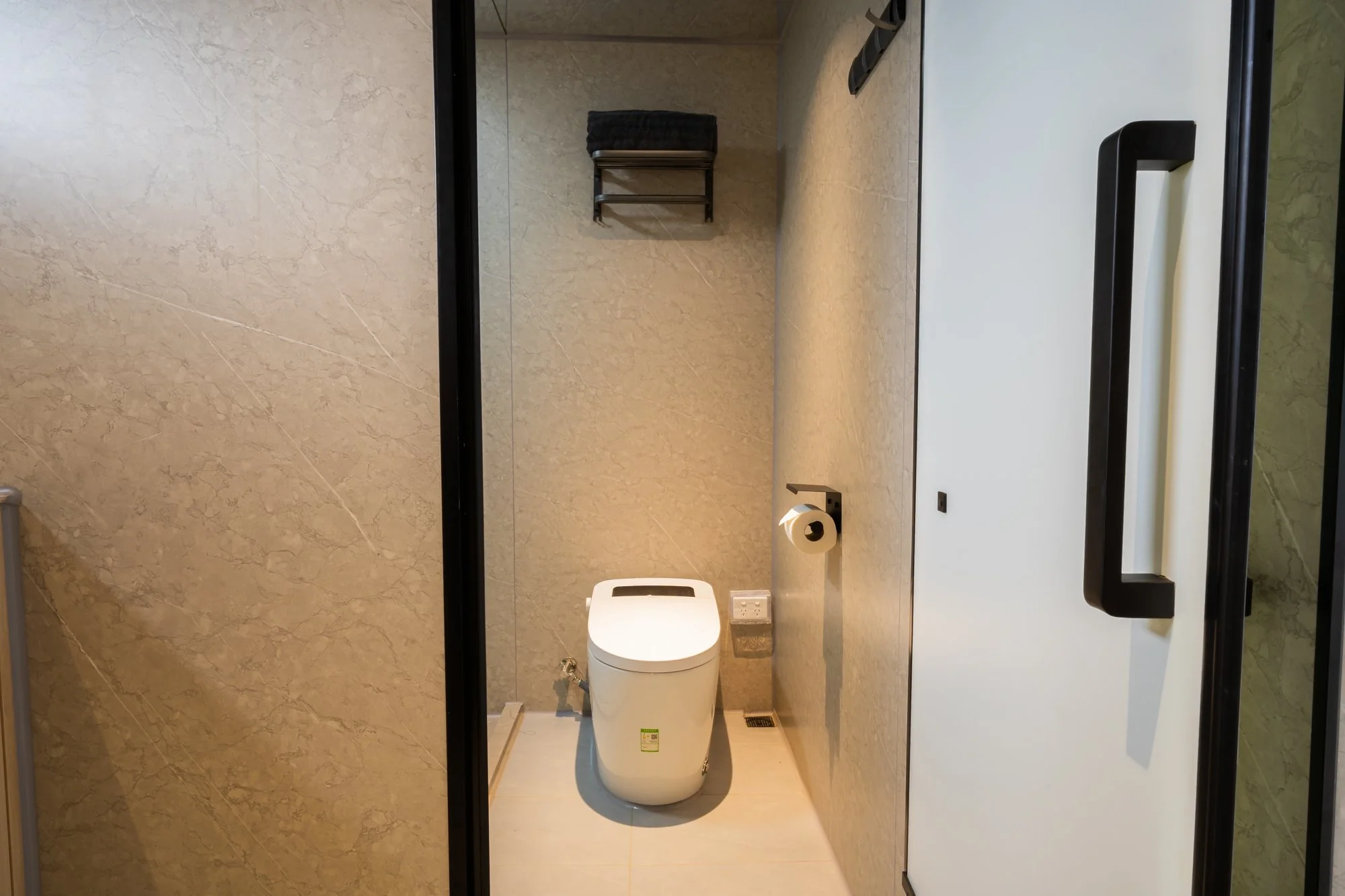 AUZPOD compact bathroom with beige marble walls, modern white toilet, black wall-mounted shelf with towel, black door handle, toilet paper holder, and floor vent.