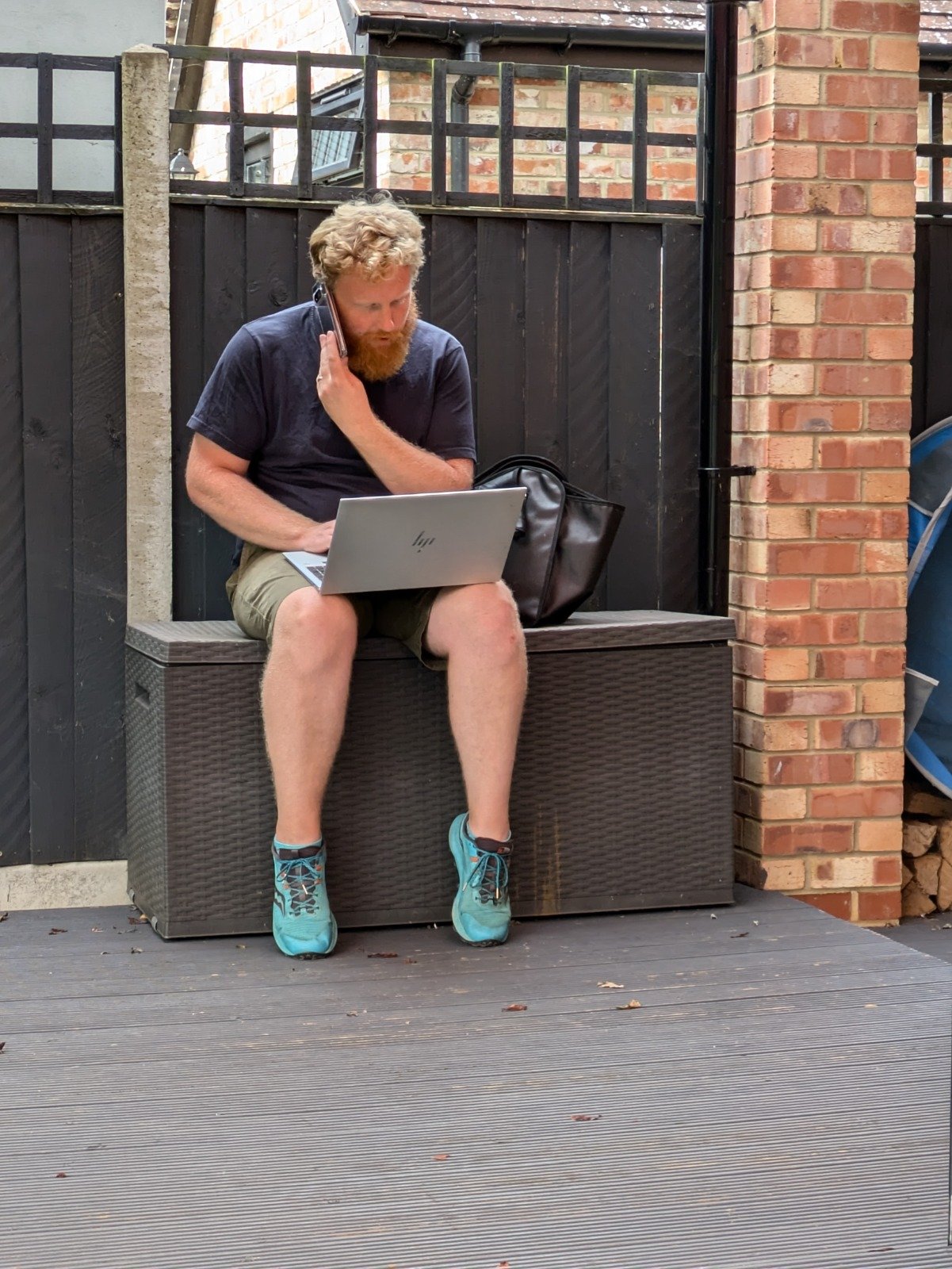 A man with curly red hair and a beard, wearing a navy t-shirt, tan shorts, and turquoise running shoes, sitting on an outdoor bench with a laptop on his lap, talking on a mobile phone, in a backyard with a wooden deck, black fence, brick wall, and a black bag beside him.