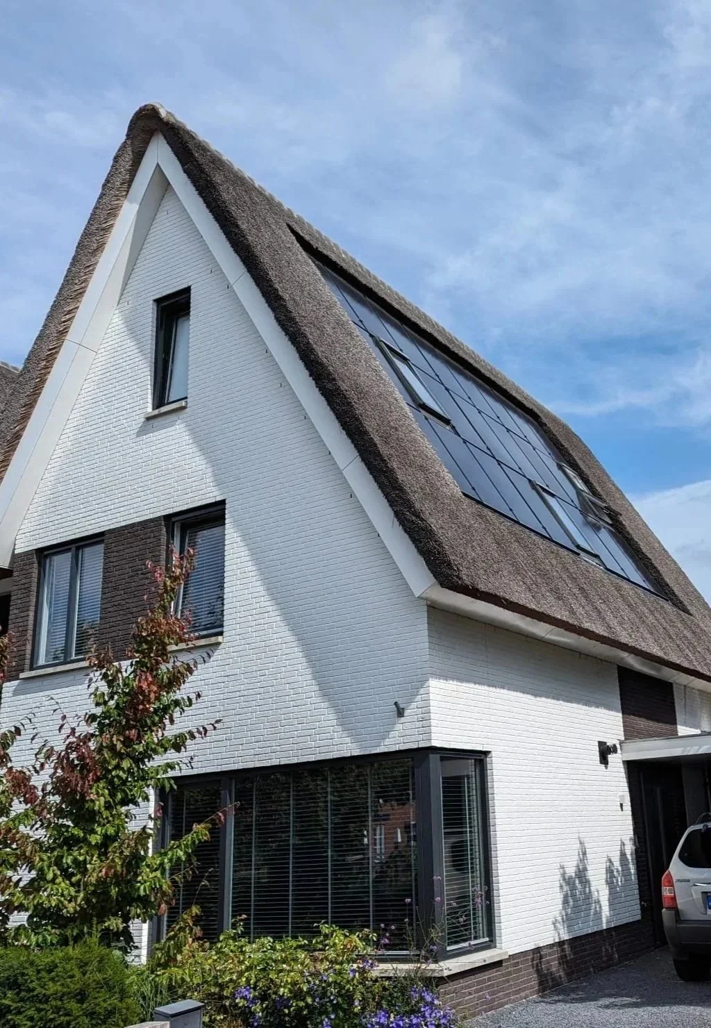 A modern house with a steeply pitched thatched roof with integrated Black solar panels, white brick exterior, large glass windows, and a small landscaped front yard with plants and a tree.