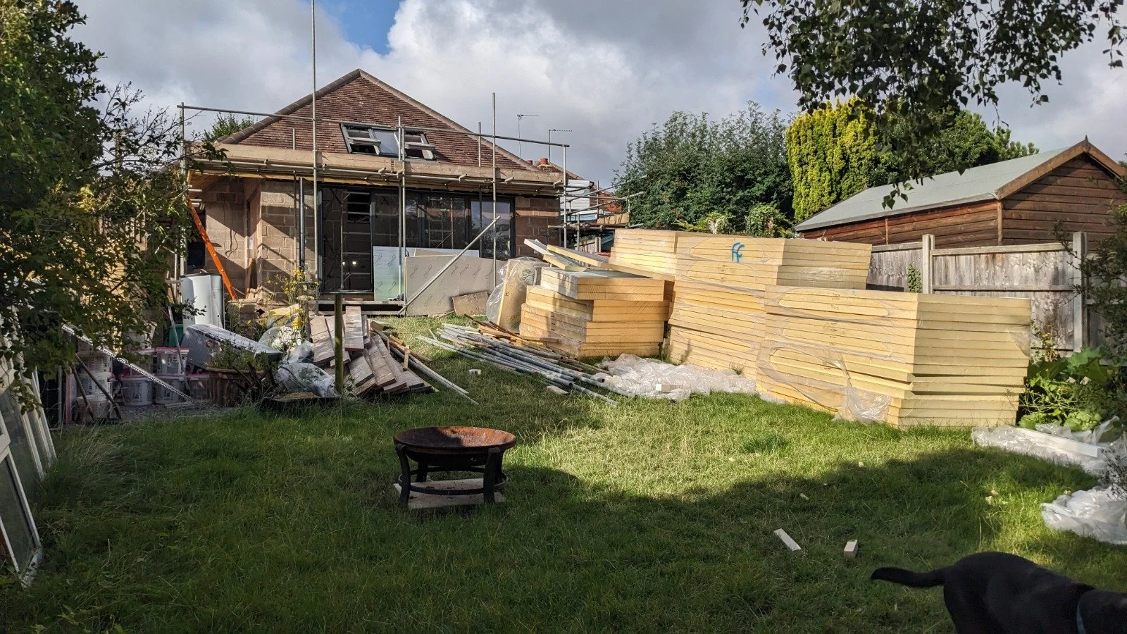 Backyard construction scene with a house under renovation, stacked lumber, construction tools and materials, and a dog near the bottom right corner.