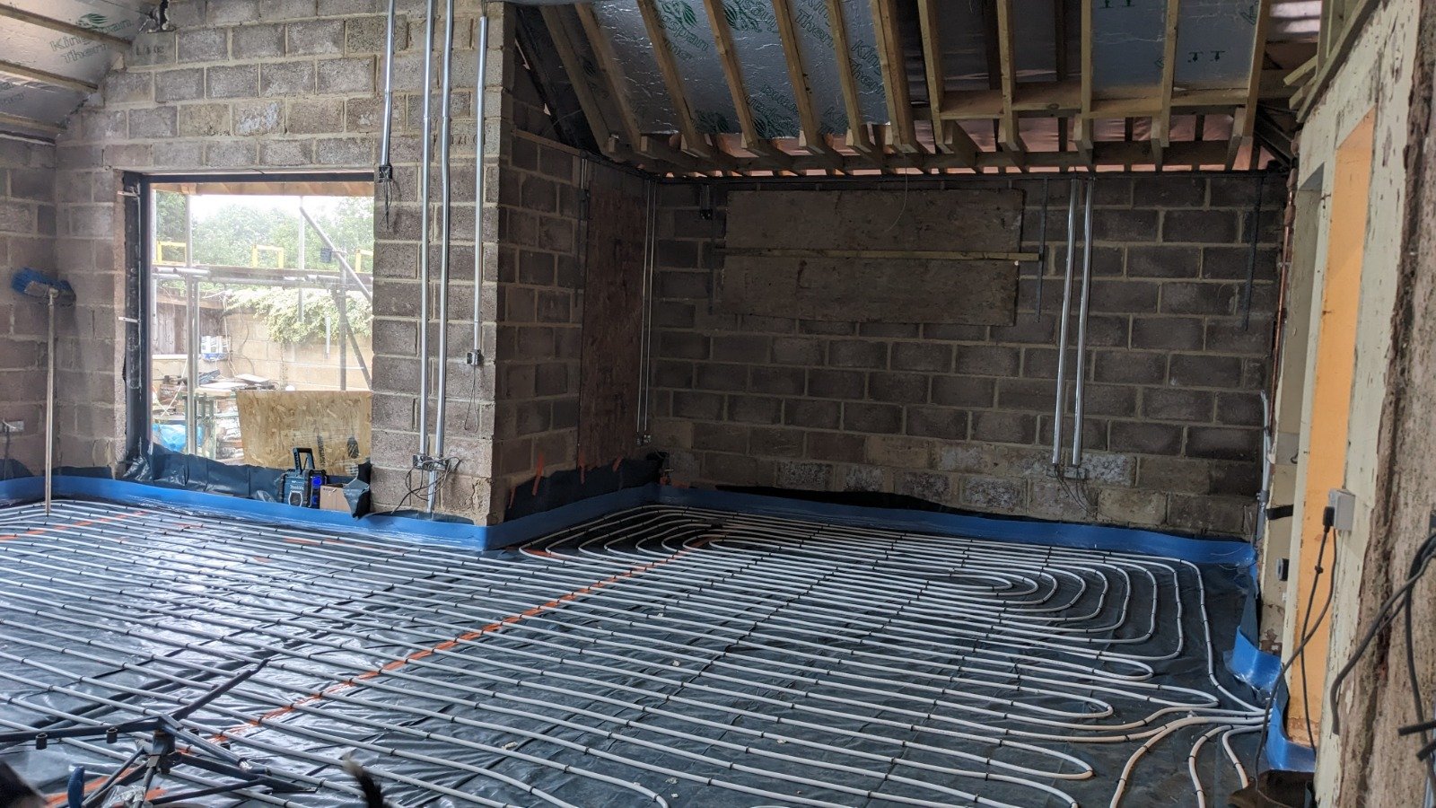 Interior of a construction site showing a radiant heating system installation with tubing laid out on the floor, partially finished walls, and an opening for a window.