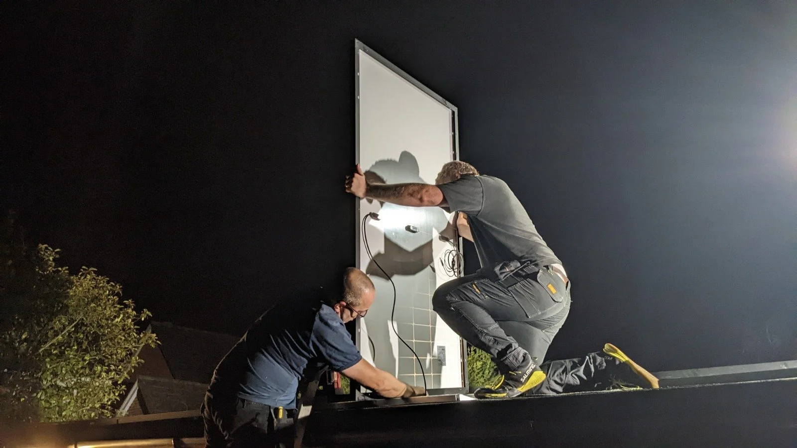 Two workers installing solar panels on a building at night. One worker is kneeling on the surface, securing the screen, while the other is standing and holding it in place.