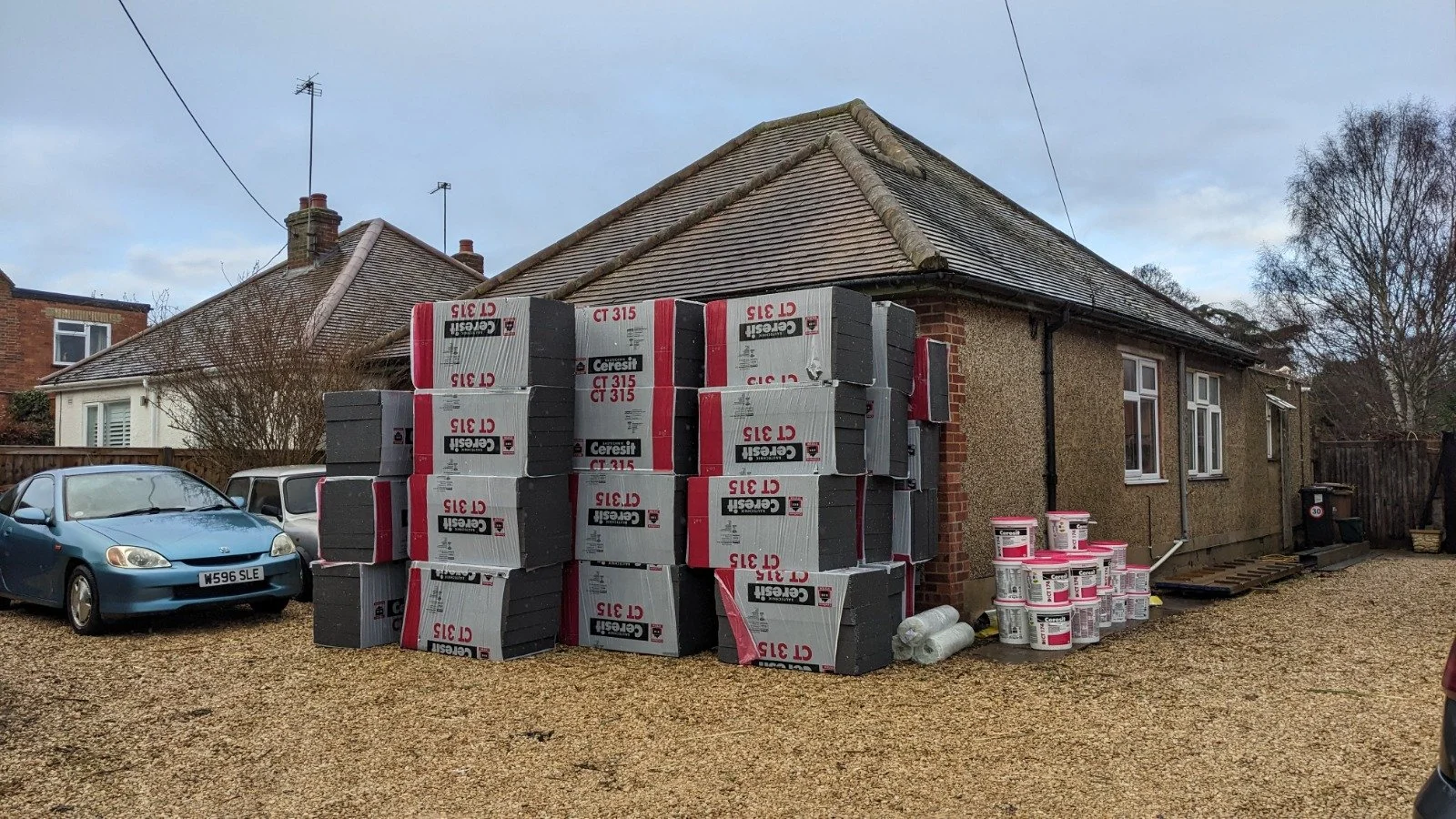 Construction materials stacked outside a house, including insulation boards and buckets of paint, on a gravel driveway.