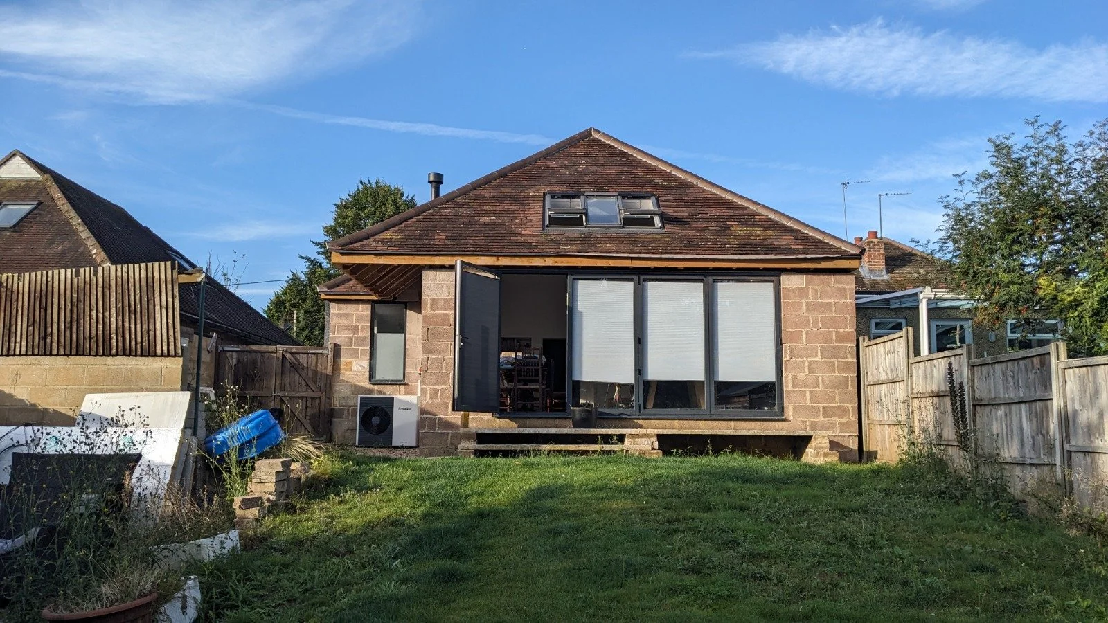 Rear view of a brick house with large sliding glass doors, open window, and a small deck; fenced backyard with green grass and some plants, blue sky with clouds.