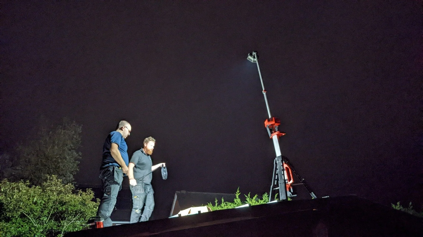 Two men installing solar panels outdoors at night with a tall extendable work light or camera boom and a work table with equipment.