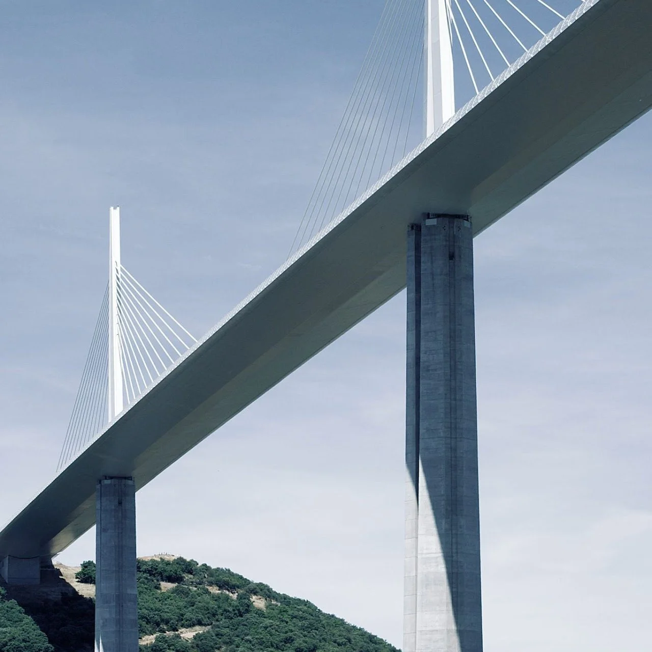 Close-up view of the San Francisco-Oakland Bay Bridge with its tall concrete pillars and white cables against a light blue sky and green hillside.