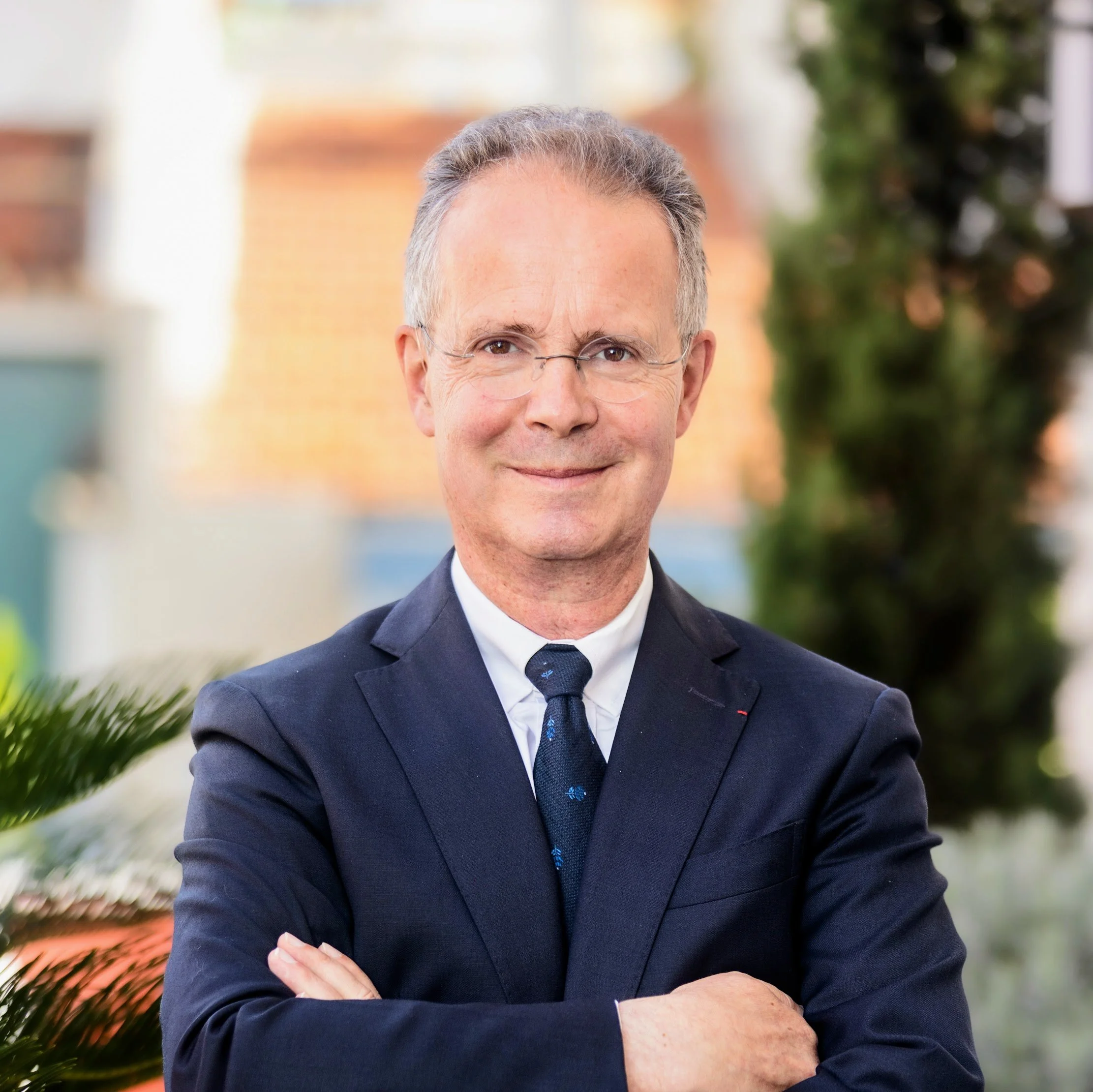 Gilles, our founder, dressed in a dark suit and tie, standing outdoors with arms crossed, smiling at the camera.