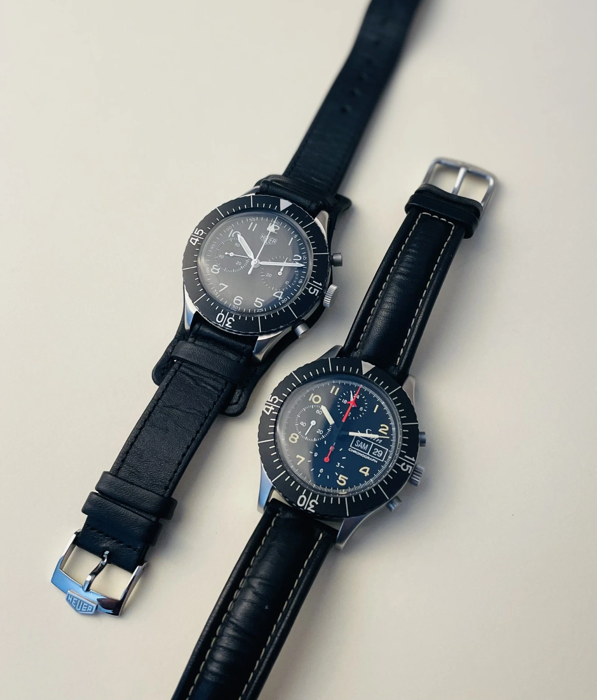 Two black leather strap chronograph watches with black bezels and dials, one branded Heuer and the other with the brand Samuel.