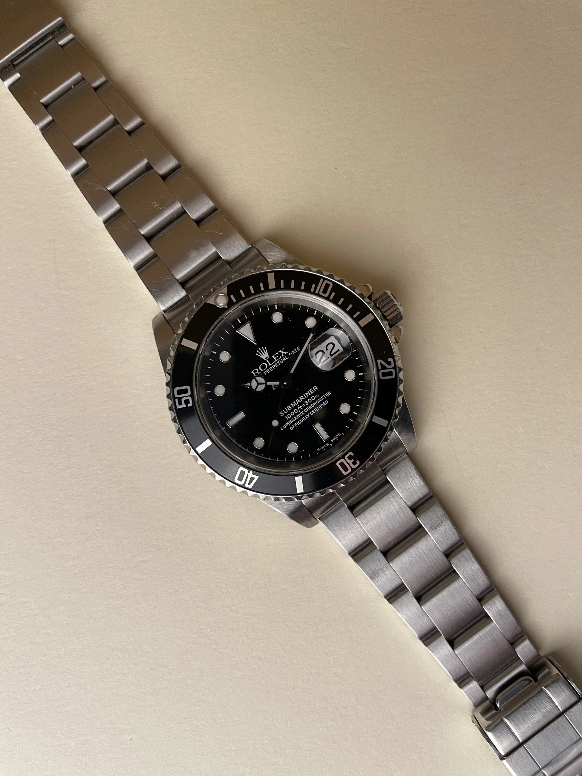 A stainless steel Rolex Submariner watch with a black dial and bezel, featuring a date window at the 3 o'clock position, on a beige background.