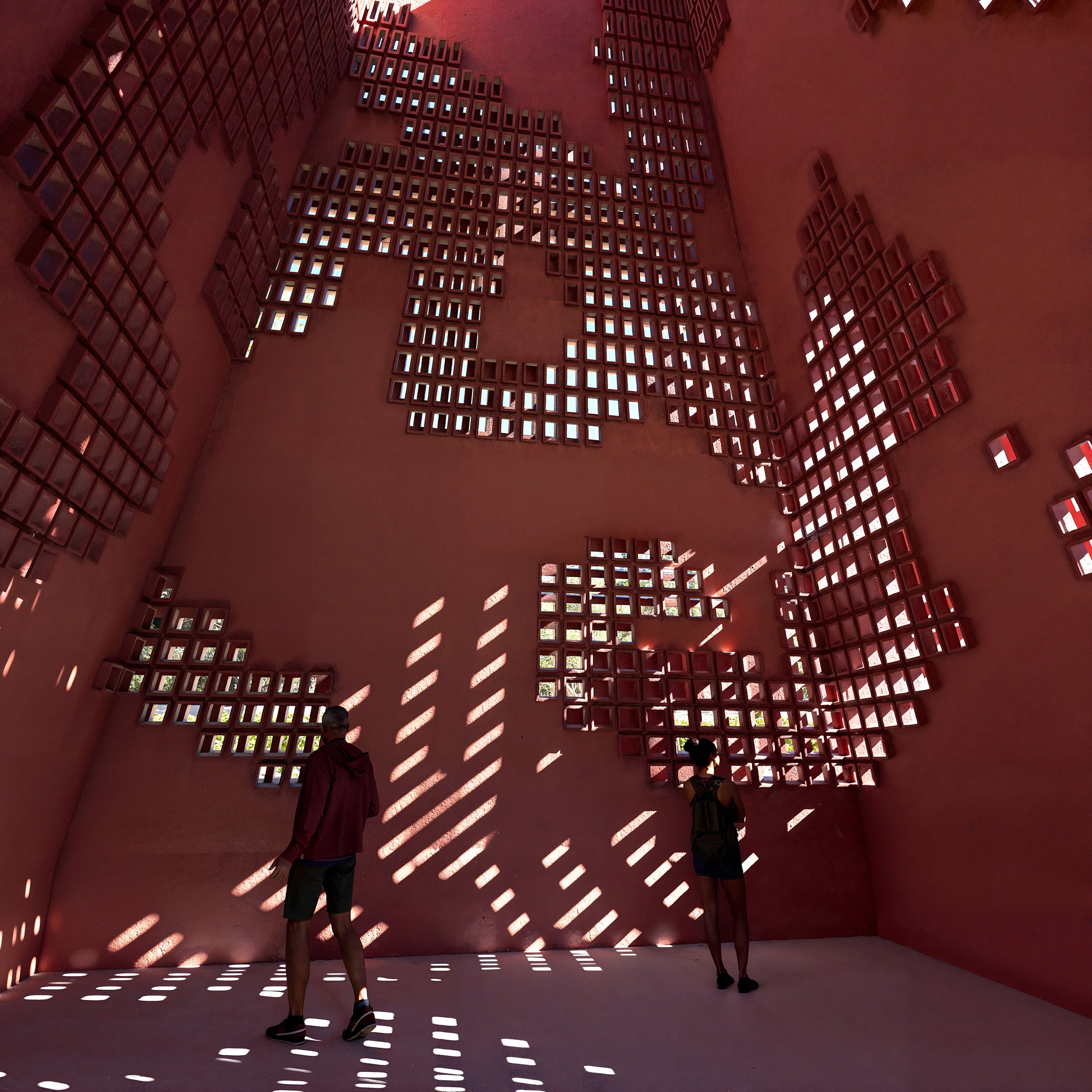 Two people standing inside a pink-colored room with a large wall featuring geometric patterned openings that cast striped shadows on the floor and walls.