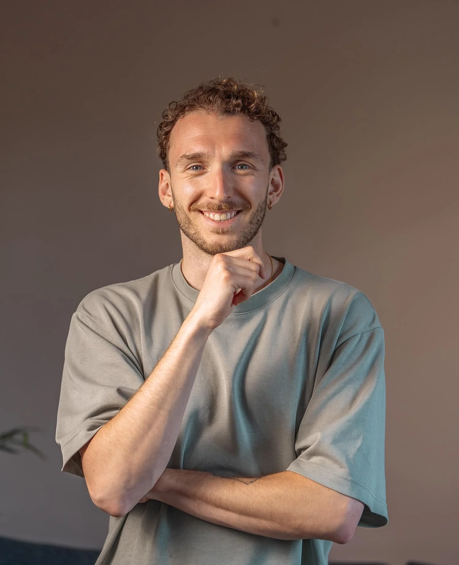A smiling young man with curly hair, beard, wearing a gray T-shirt, standing against a neutral background with one hand resting under his chin and the other arm crossed.