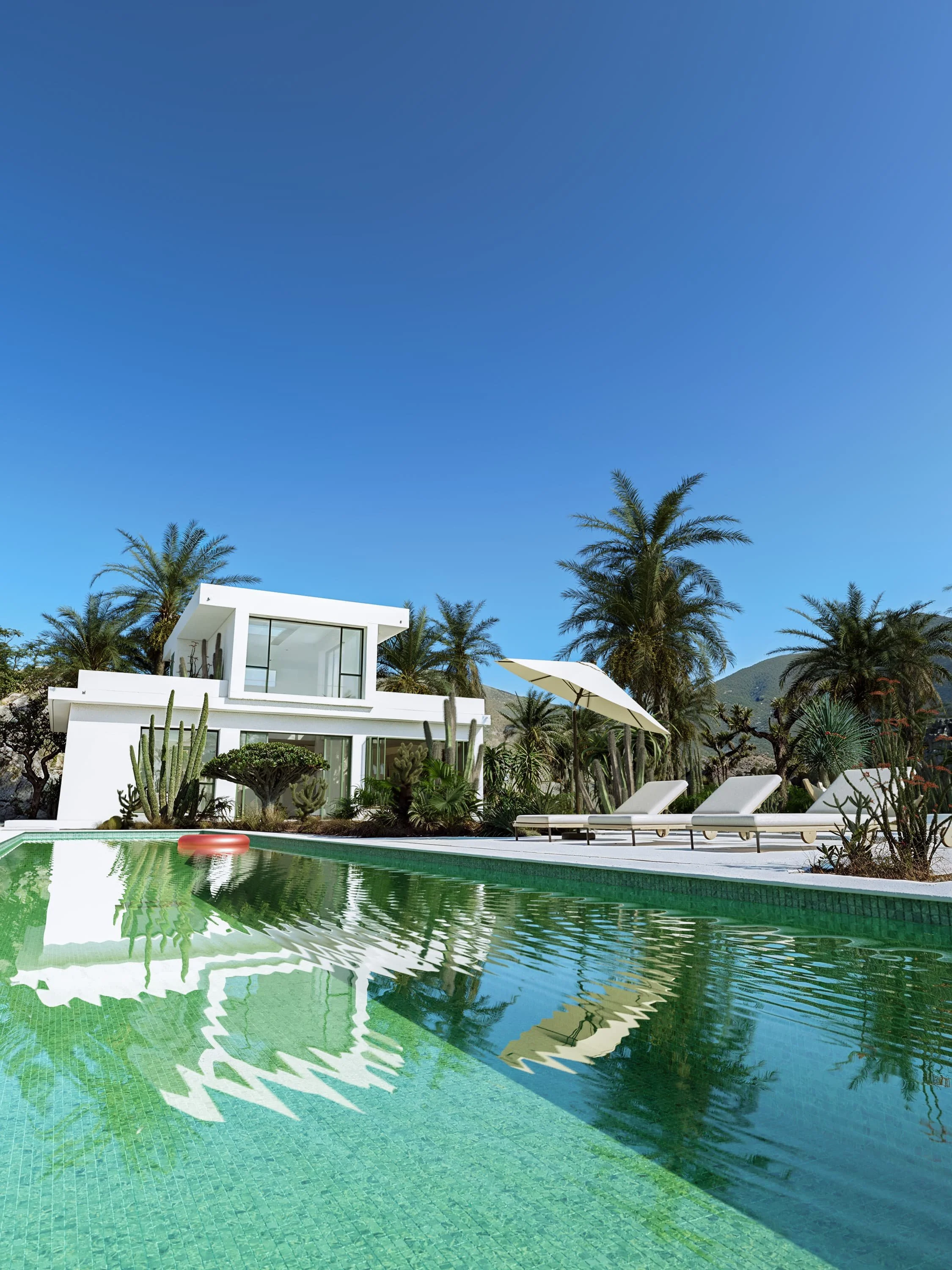 Modern white house with large glass windows, pool with green water, lounge chairs, white umbrella, and palm trees under a clear blue sky.