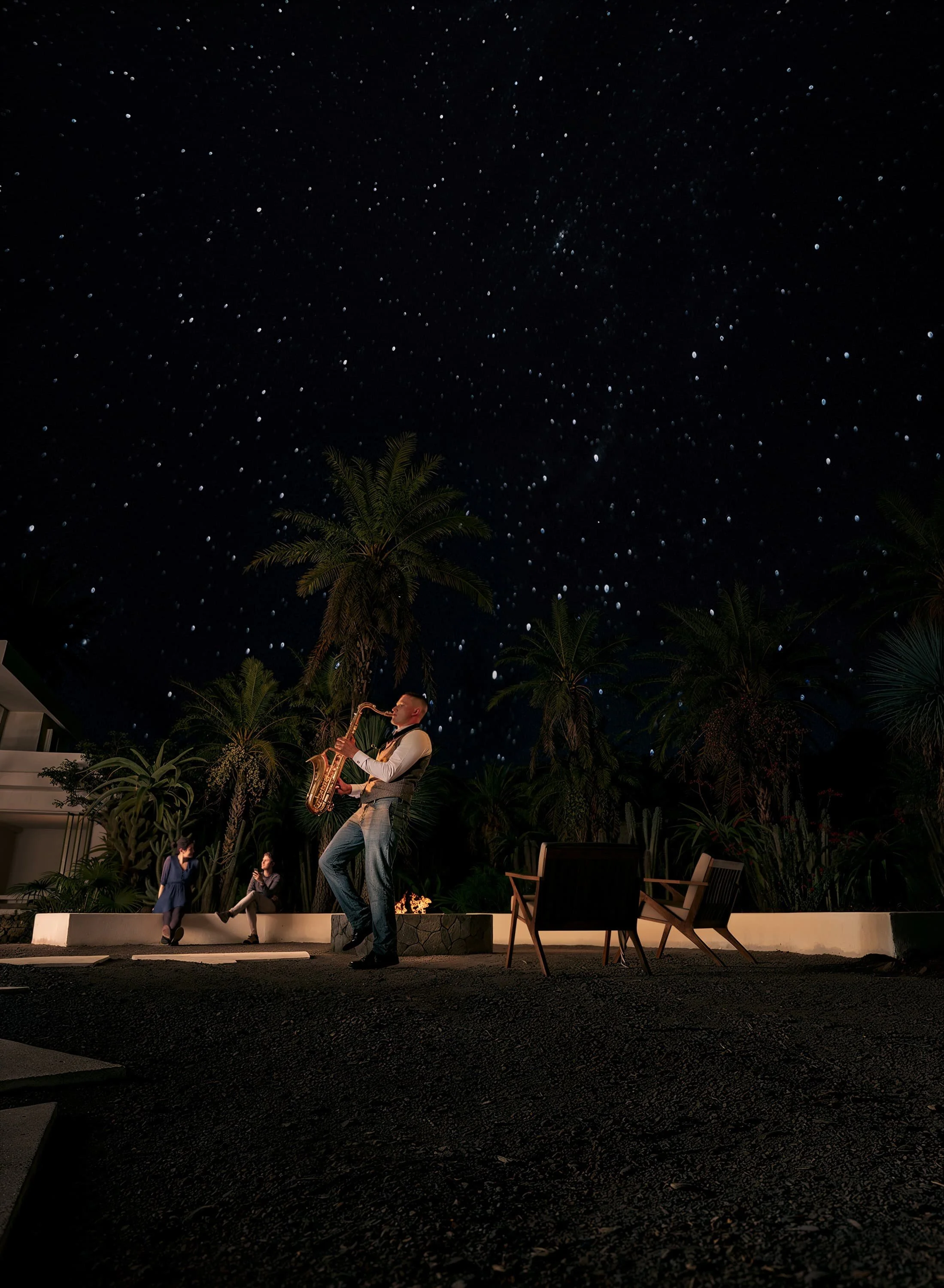 A man playing saxophone outdoors at night with children watching under a starry sky and palm trees.