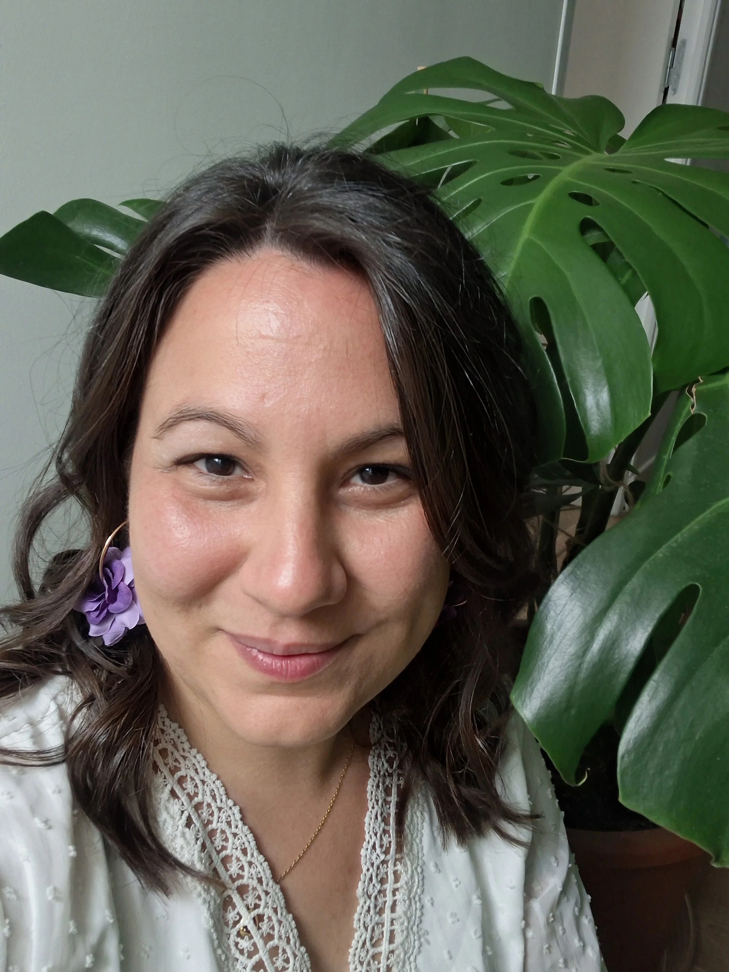 Une femme souriante avec des boucles d'oreilles violettes, portant un haut blanc à dentelle, posant près d'une plante verte avec de grandes feuilles.
