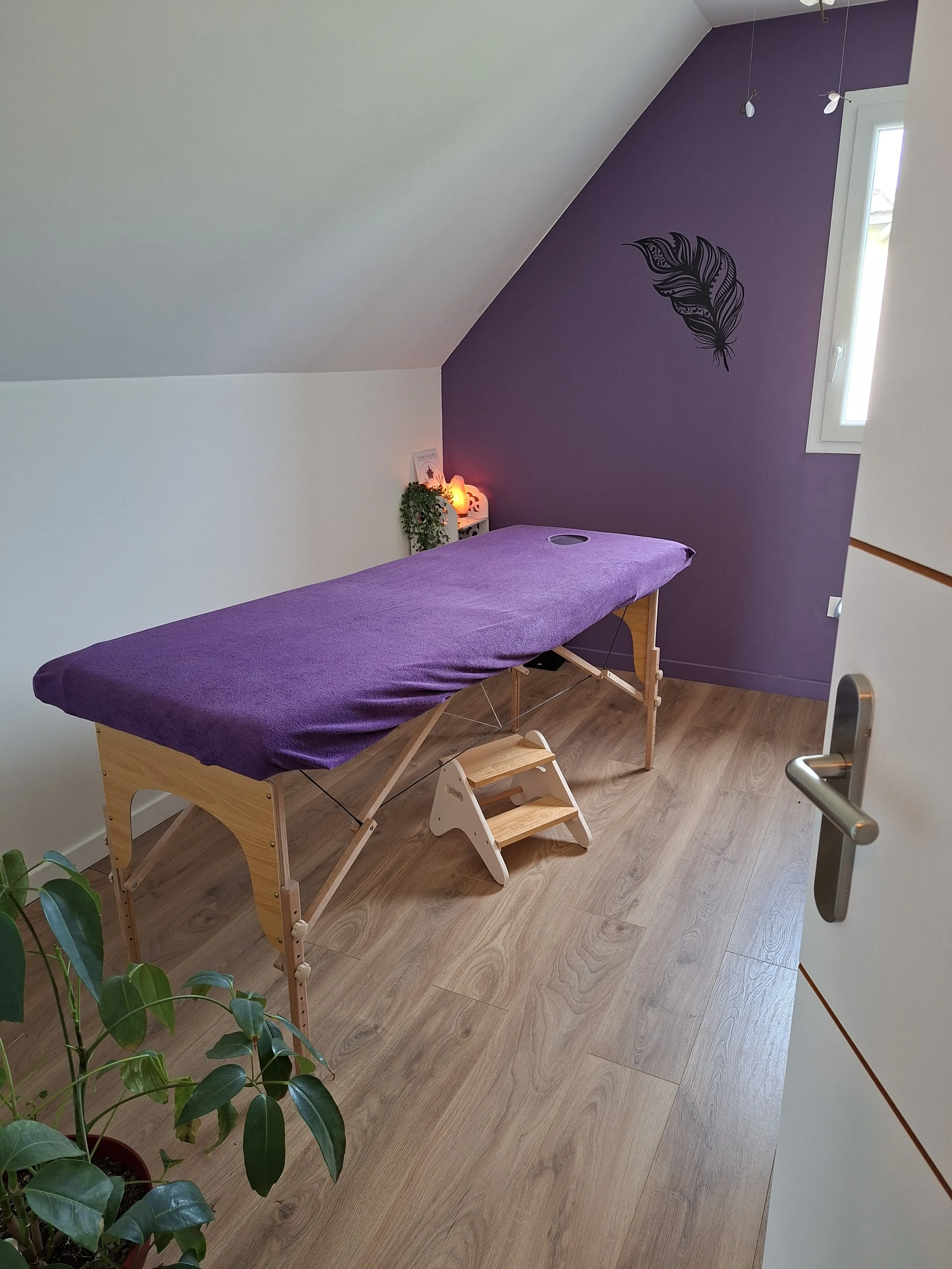 Une salle de massage avec une table recouverte d'une couverture violette, des murs blanc et violet avec un motif de plume, une petite étagère à côté avec une plante et une lampe orange, un rideau blanc en haut et un sol en bois.