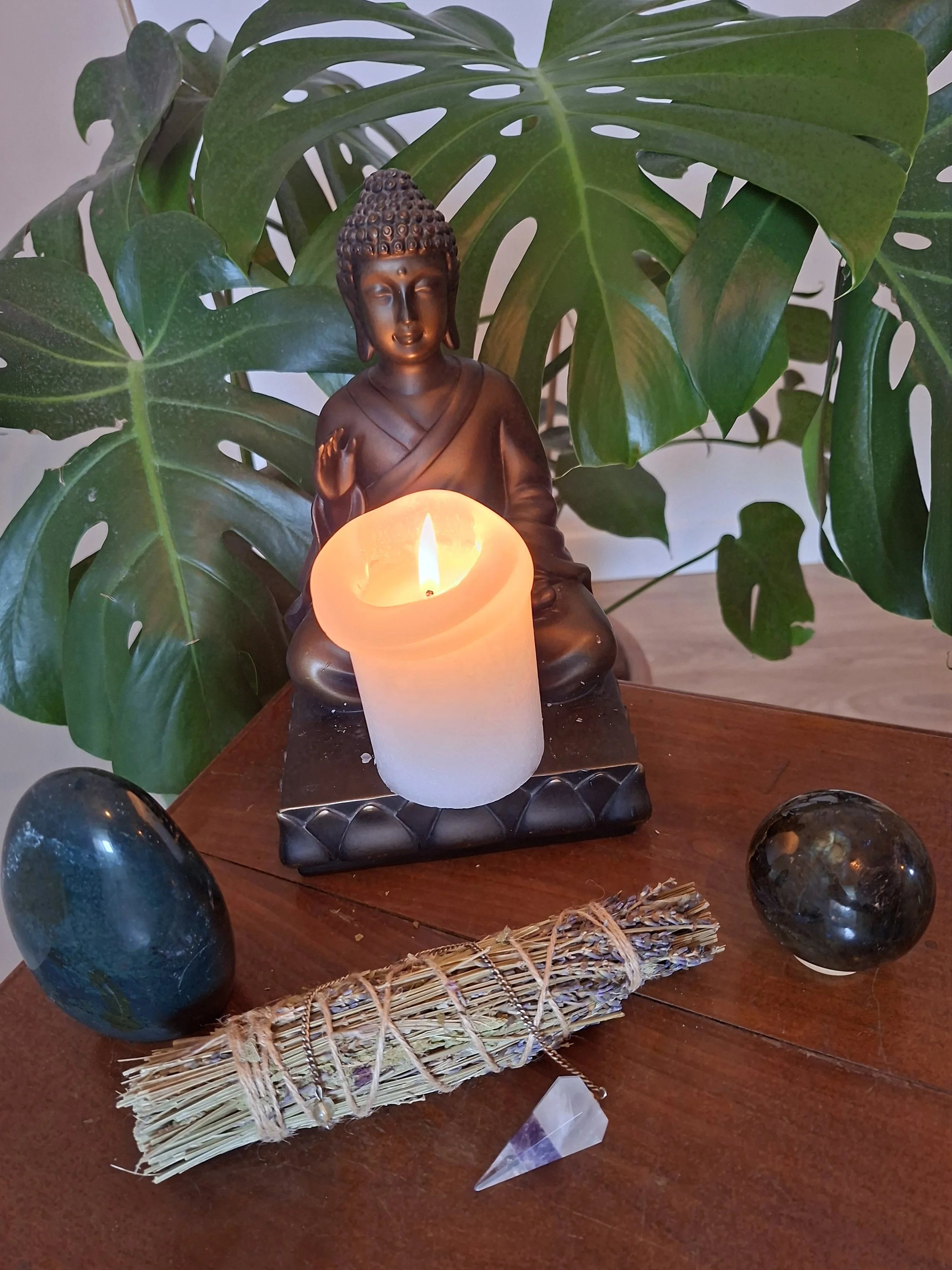 Une statue de Bouddha avec une bougie allumée devant, entourée de deux cristaux, une pierre noire et une pierre verte, avec une plante verte en arrière-plan.