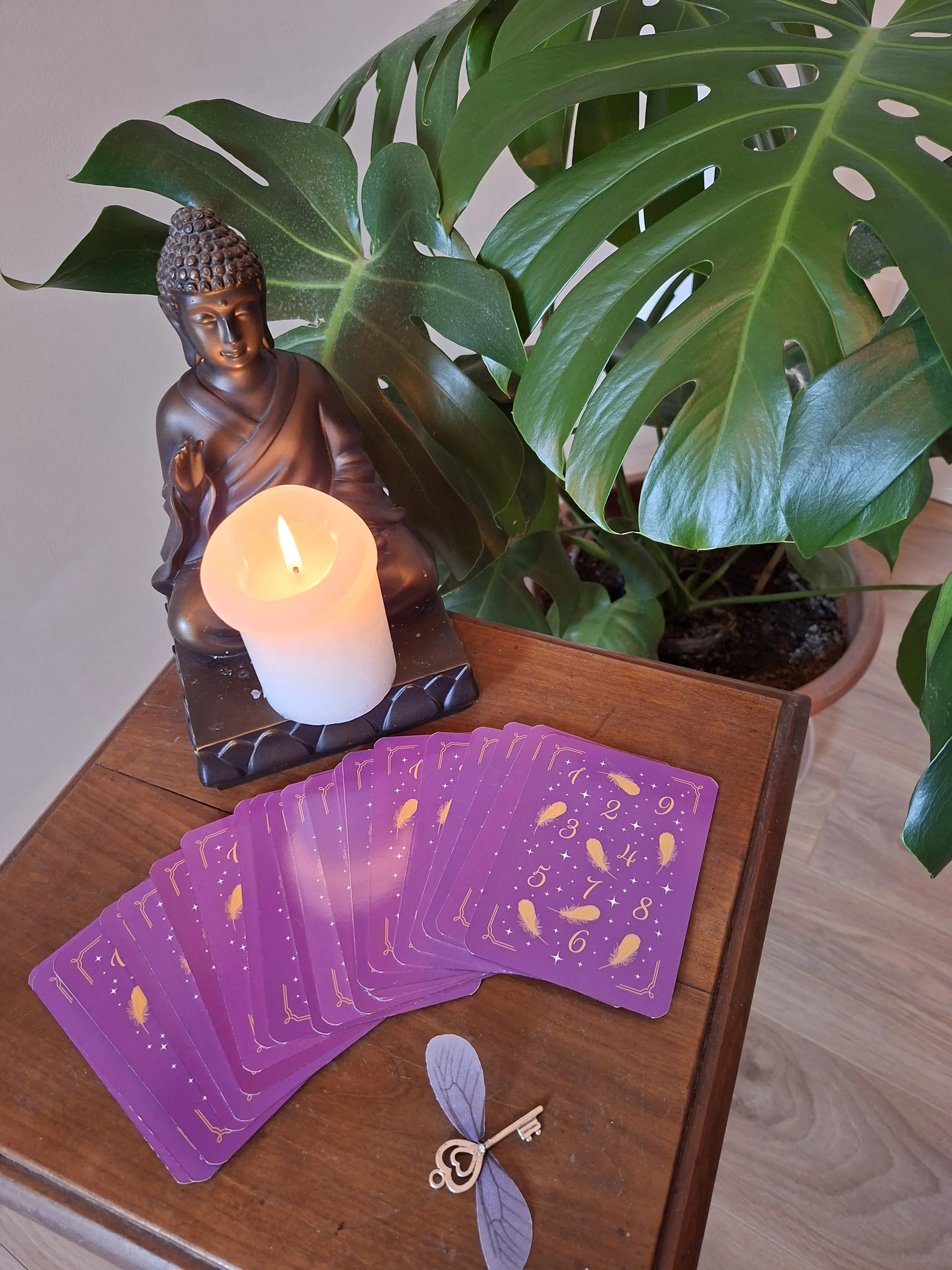 Jeu de tarot avec cartes violettes, figurine de Bouddha, bougie allumée, petite clé en forme d'insecte, et grande plante verte dans un pot en terre cuite sur une table en bois.