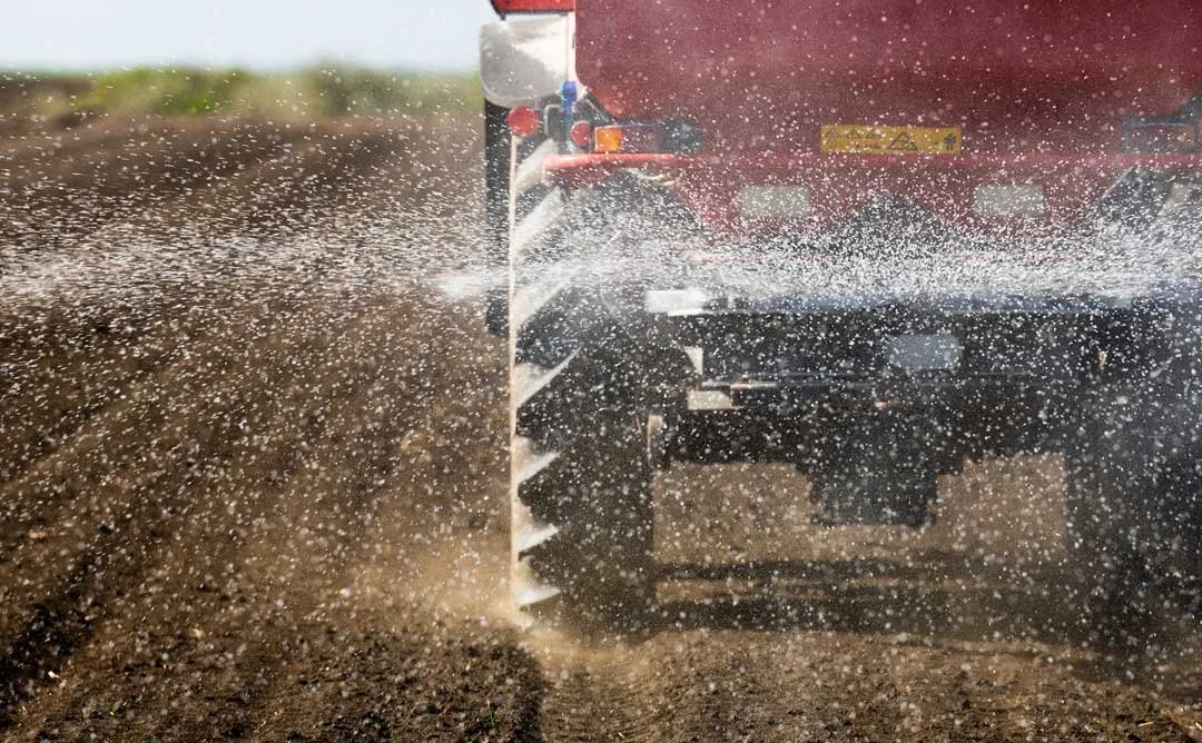 Close-up of a tractor spreading fertilizer or seeds on a farm field, with dust and particles flying in the air.
